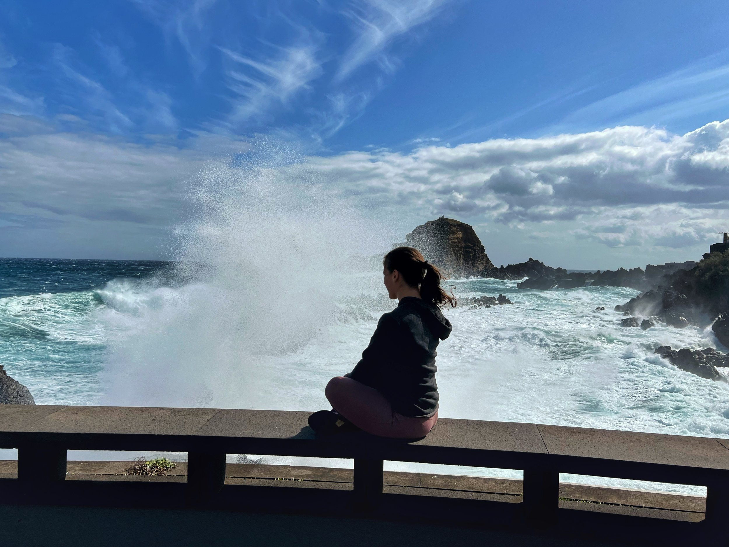 A person sitting on a bench near the ocean, gazing at the waves splashing against rocks with a large rock formation in the background under a partly cloudy sky.
