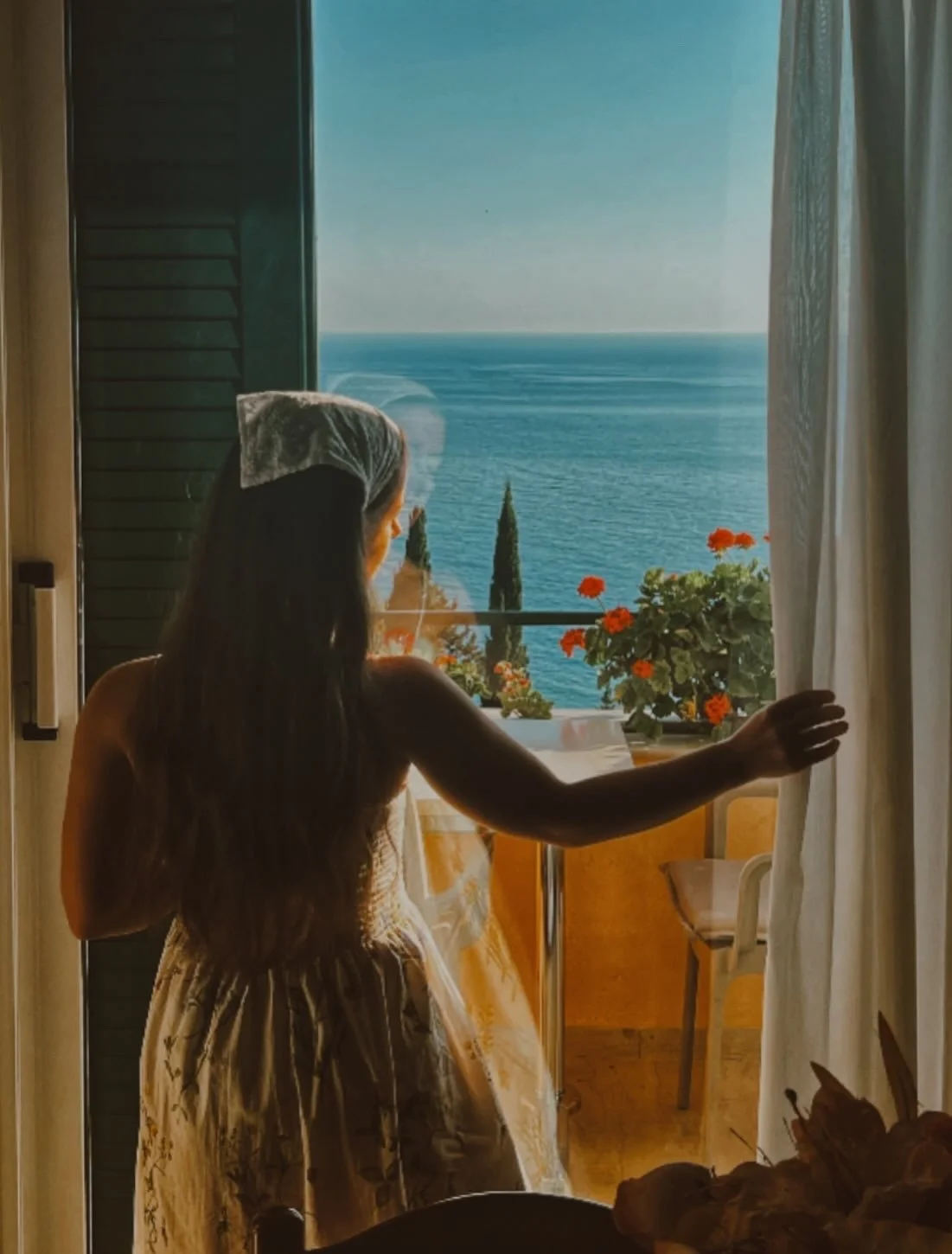A woman with long hair, wearing a lace head covering and a dress, stands indoors by a window with closed curtains, looking out at a view of the ocean, with trees and flowers in the foreground.