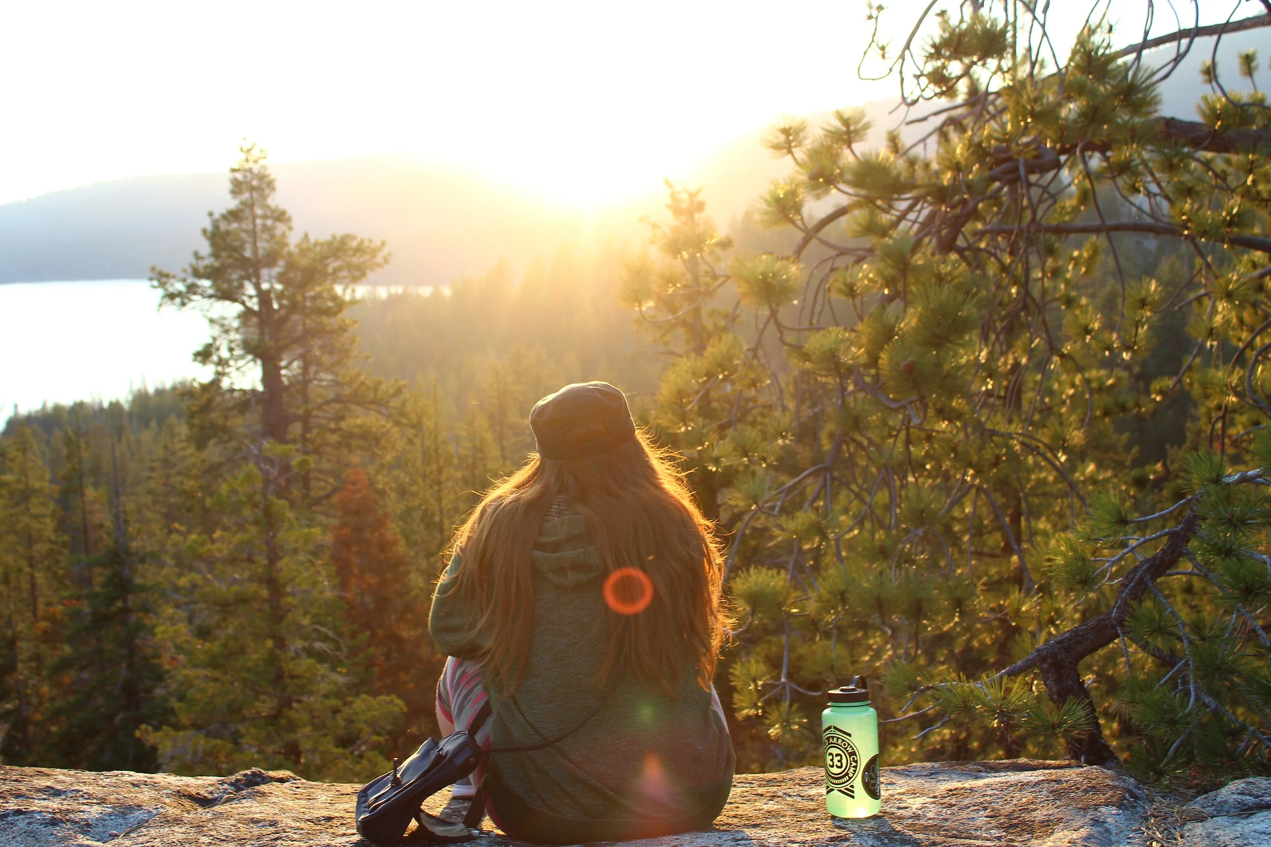 A woman with long red hair sitting on a rock in a forest at sunset, overlooking trees and mountains, with a water bottle beside her.