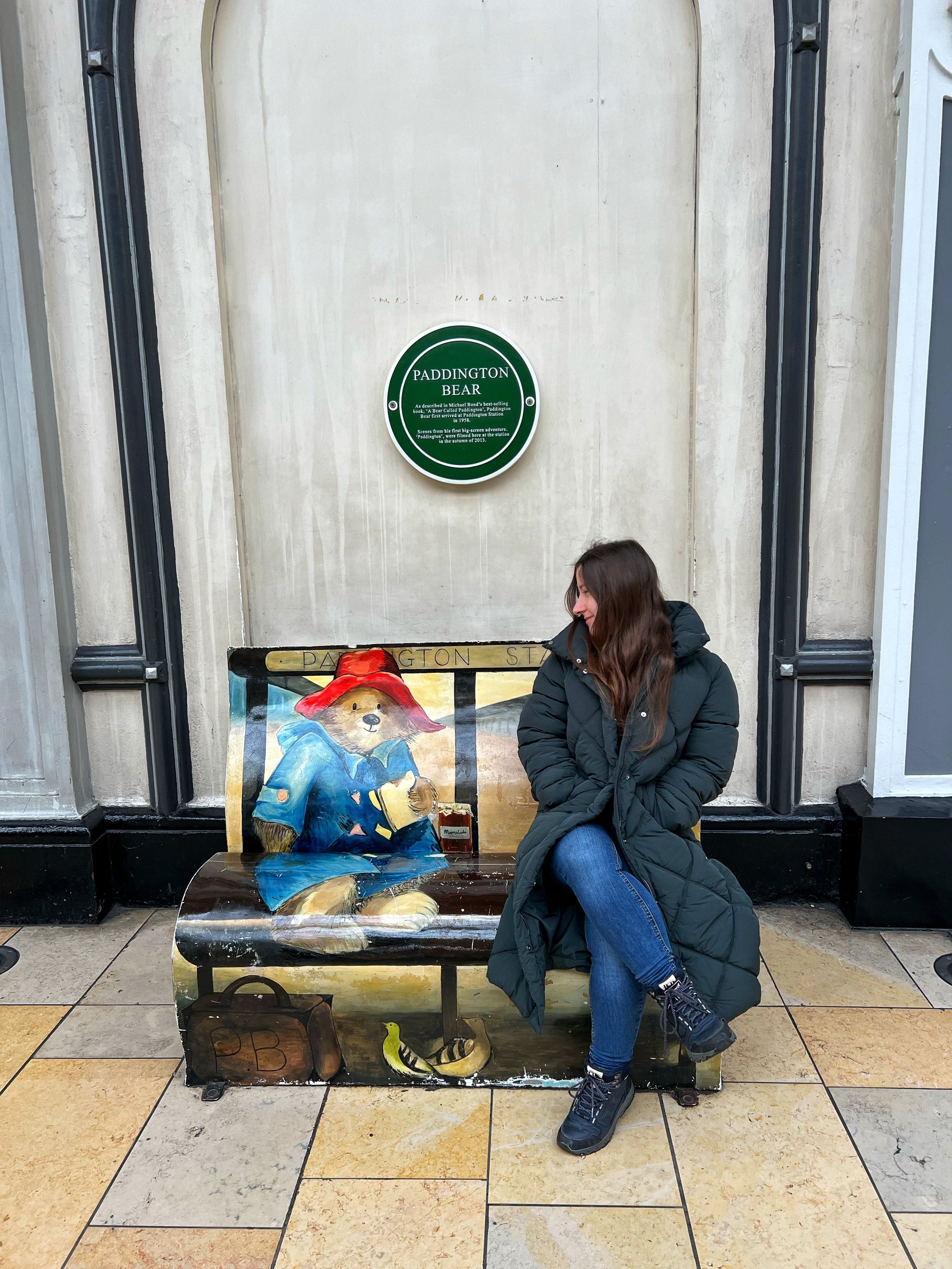 A woman with long brown hair, wearing a black puffer jacket, blue jeans, and black outdoor boots, sitting on a painted wooden bench with her arms crossed and smiling, in front of a mural of Paddington Bear at Paddington Station.