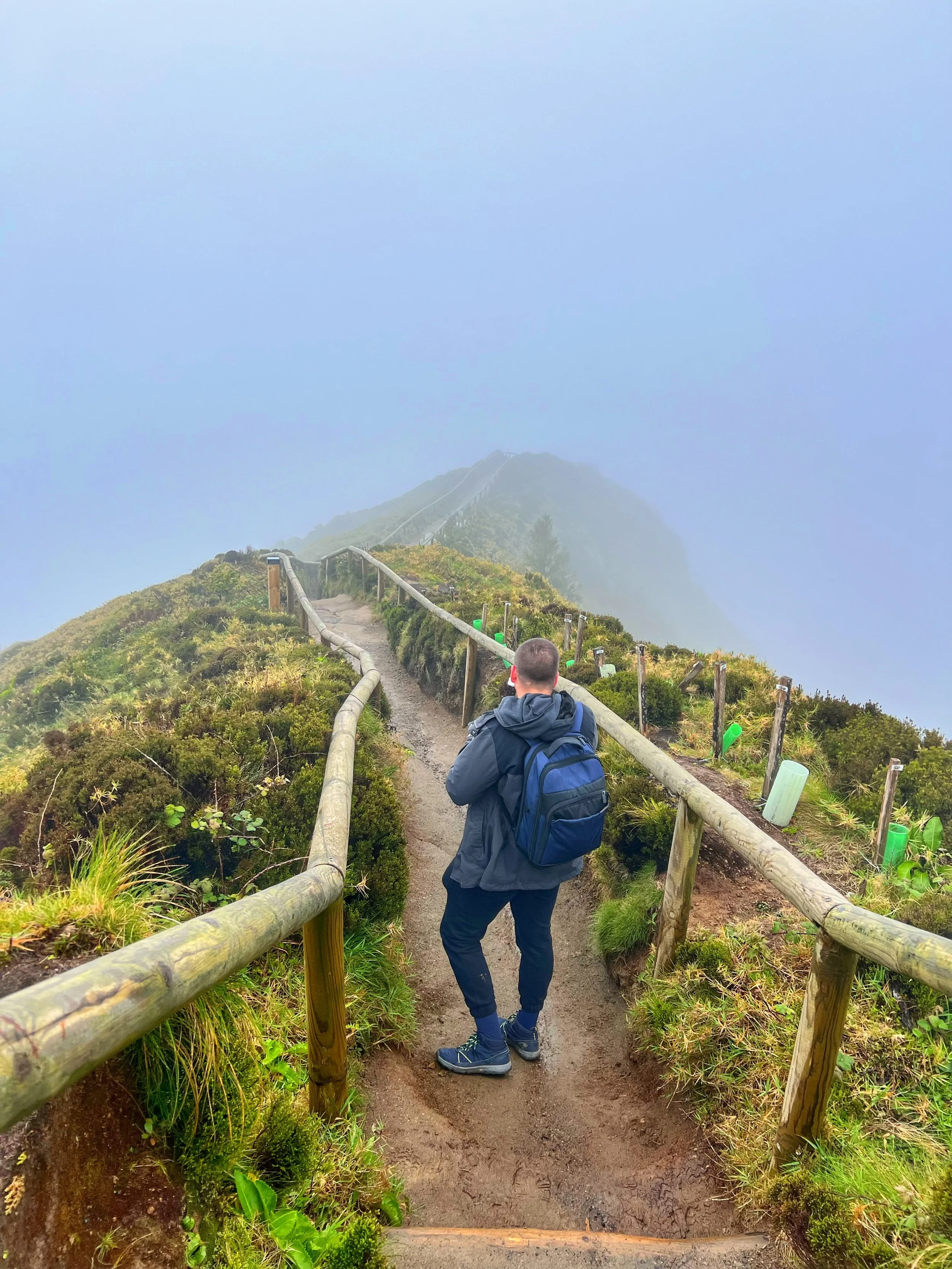 A person walking on a narrow mountain trail with a wooden fence on both sides, surrounded by green plants and shrubs, leading towards a misty, foggy mountain peak.