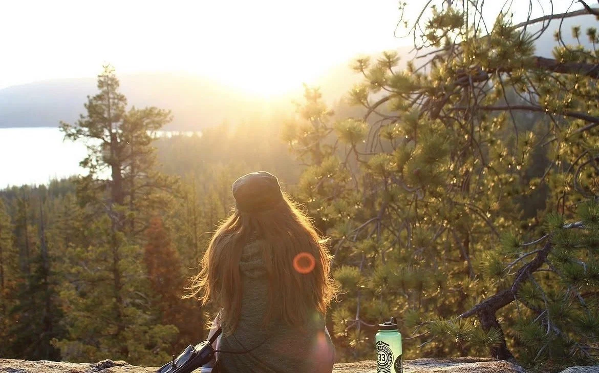 A woman with long hair sitting on a rock, watching a sunset over a forested landscape with pine trees. She has a water bottle and a camera beside her.