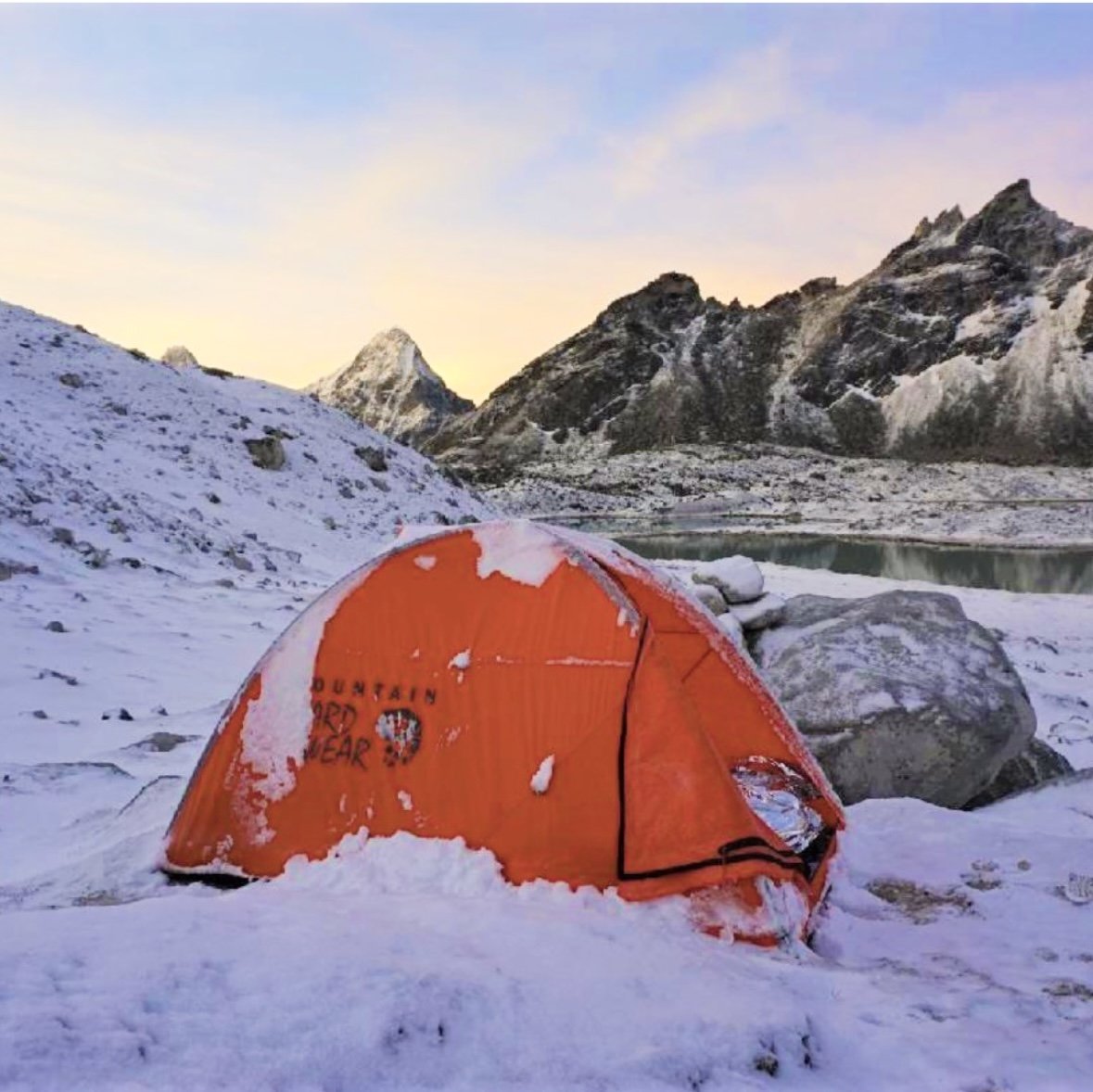 Orange tent set up on snow-covered ground in a mountainous landscape with snow-capped peaks and a lake in the background at sunset. Cho-Oyu, Nepal, Gokyo, Everest Base Camp Trek.