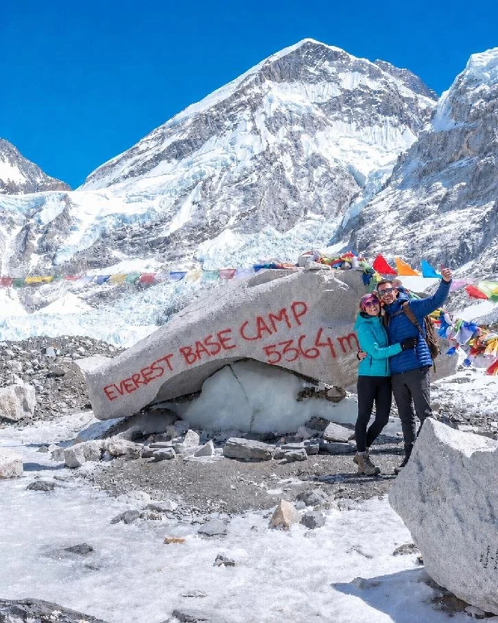 Two people in outdoor gear standing and smiling near a large boulder at Everest Base Camp, 5364 meters above sea level, with snow-covered mountains and prayer flags in the background.