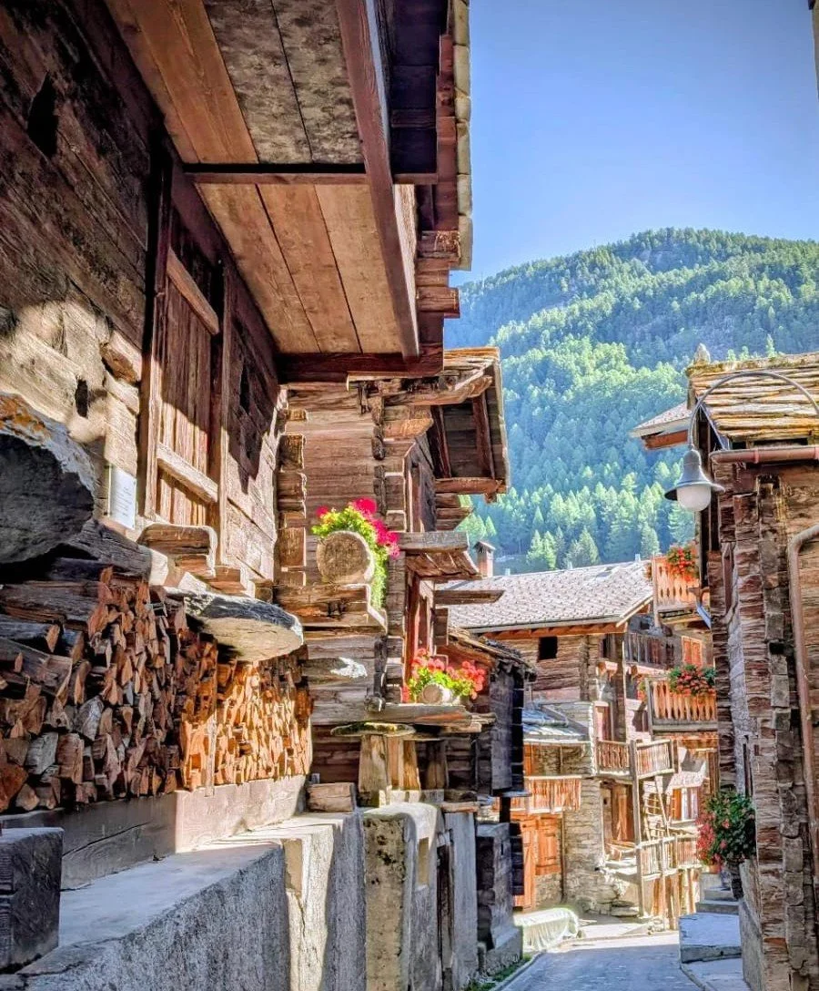 A narrow mountain village street with wooden and stone buildings, some with flower boxes, and a forested hillside in the background under a blue sky. Zermatt.