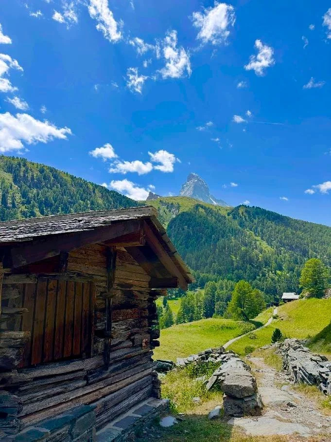 A rustic wooden cabin in a green mountainous landscape with a dirt path, under a bright blue sky with fluffy white clouds. Zmutt, Zermatt, Culturweg.