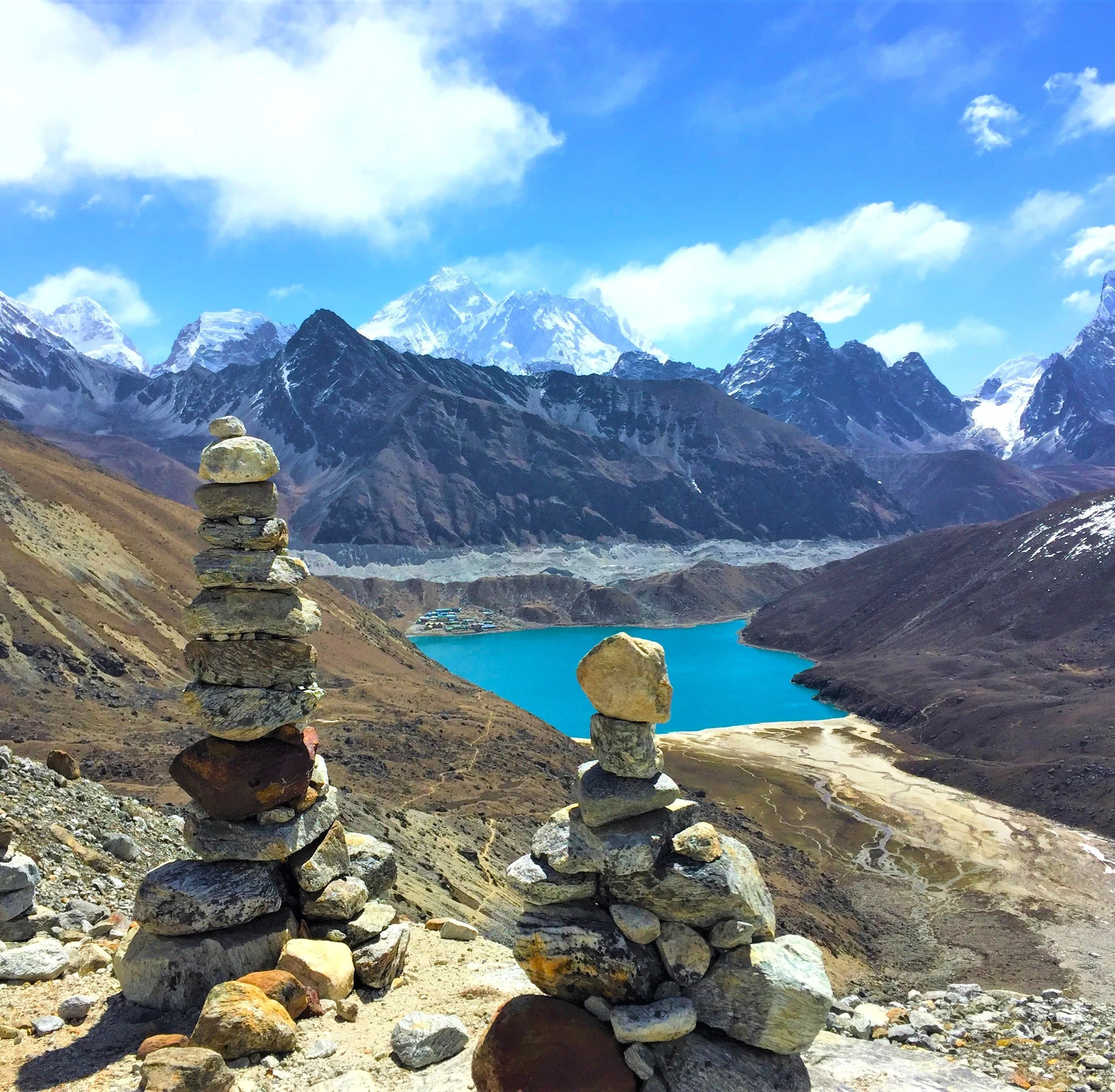 Stacked rocks in front of a mountain range with snow-capped peaks and a blue Gokyo lake, Nepal, under a partly cloudy sky.