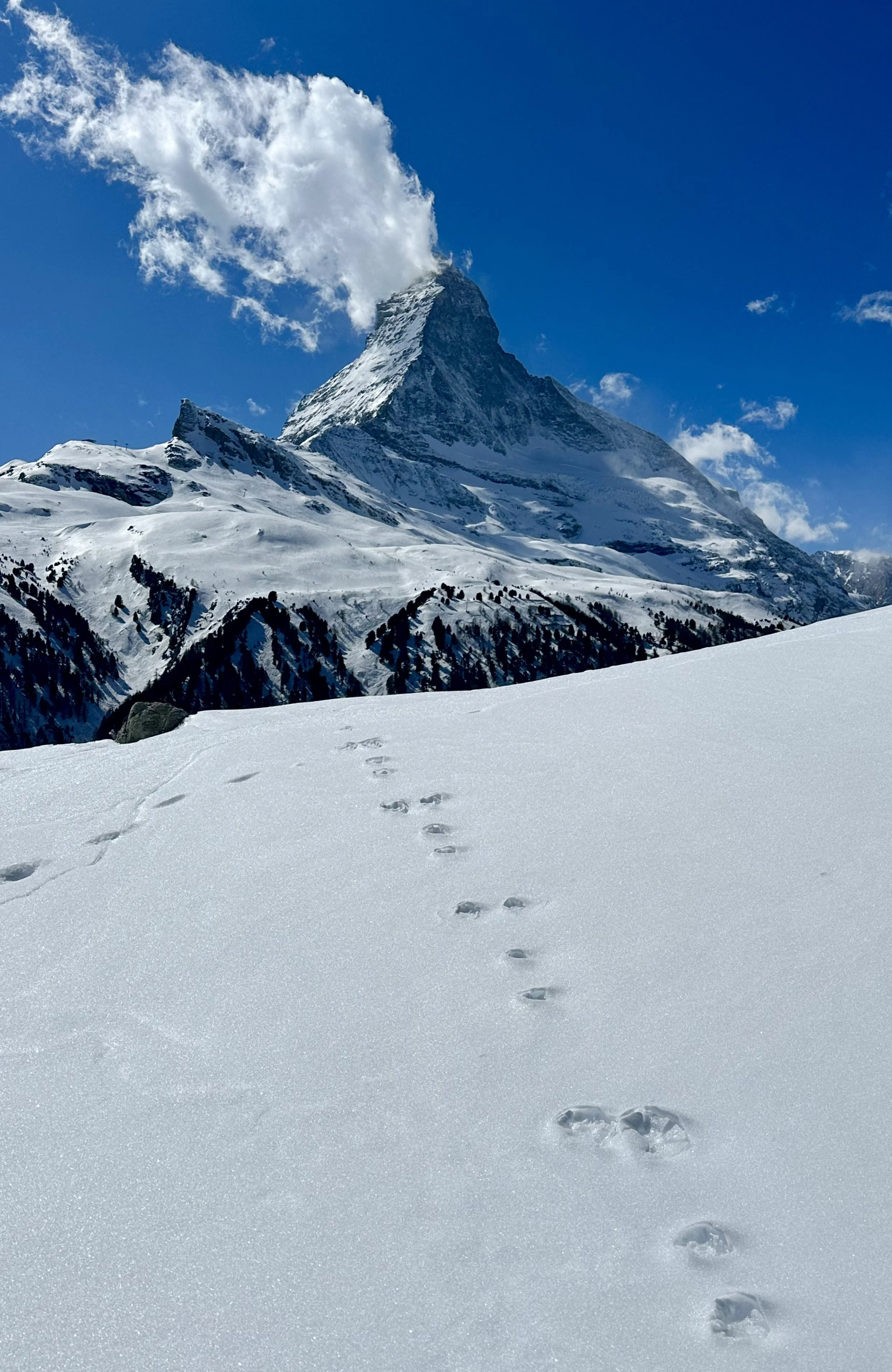 Snow-covered mountains with footprints in the snow in the foreground, and Matterhorn with clouds emitting from its summit in the background.