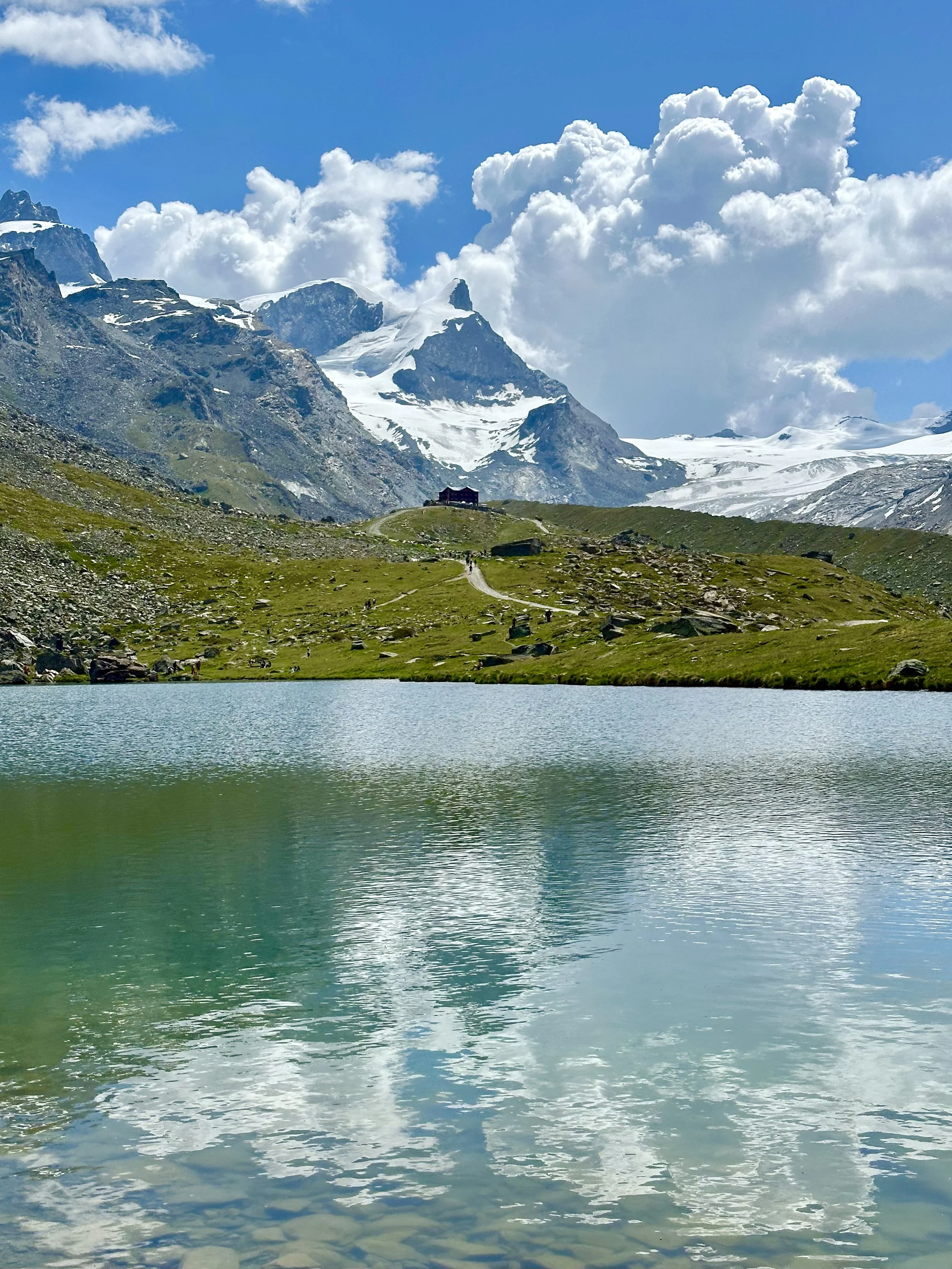 A scenic view of a mountain lake with clear, green water, a green grassy shoreline, and snow-capped mountains in the background under a blue sky with white, fluffy clouds. Five lakes hike, Zermatt, Matterhorn.