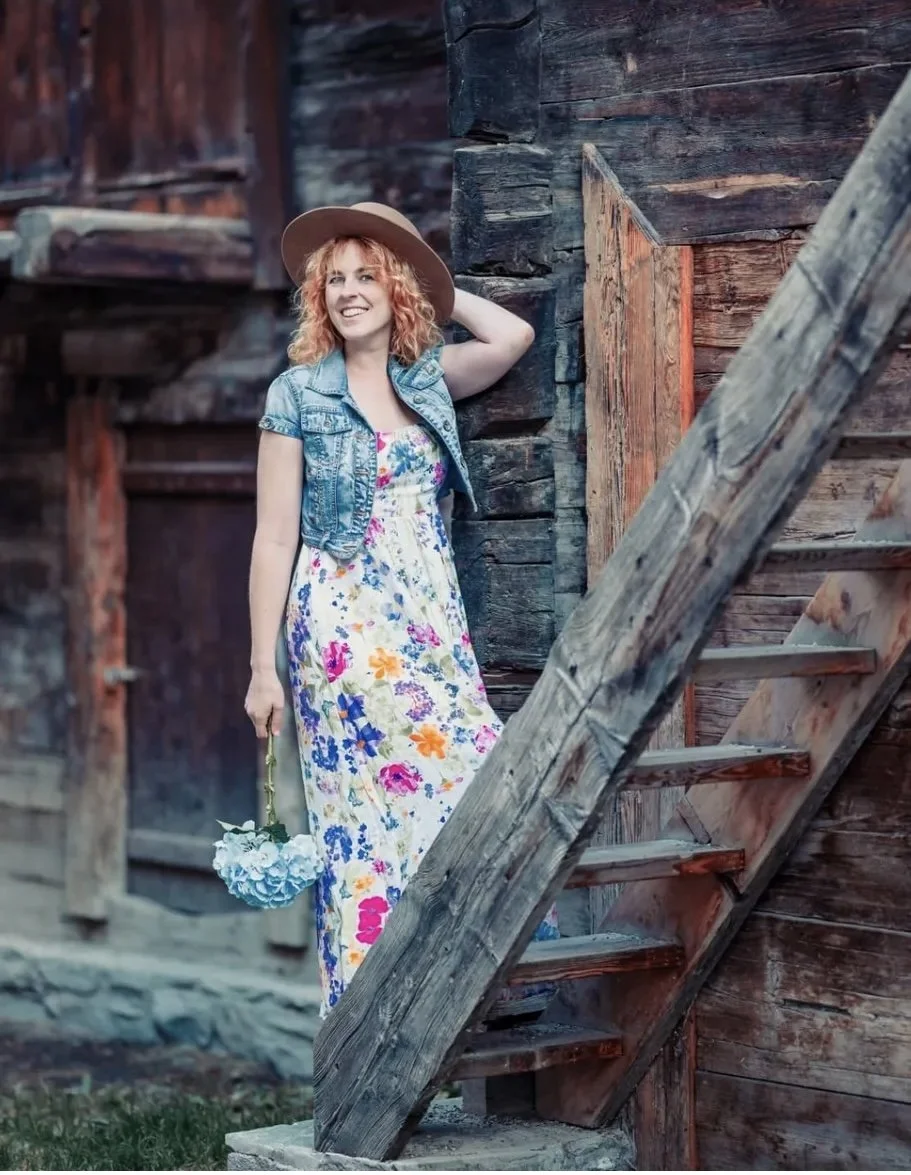 Woman with curly red hair wearing a wide-brimmed hat, denim vest, and a colorful floral dress, holding a bouquet of white flowers, standing beside an old wooden building with rustic stairs.