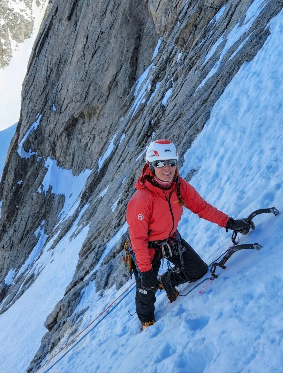 A female climber in a red jacket with climbing gear, helmet, and sunglasses ascending a snowy mountain slope near a rocky cliff.