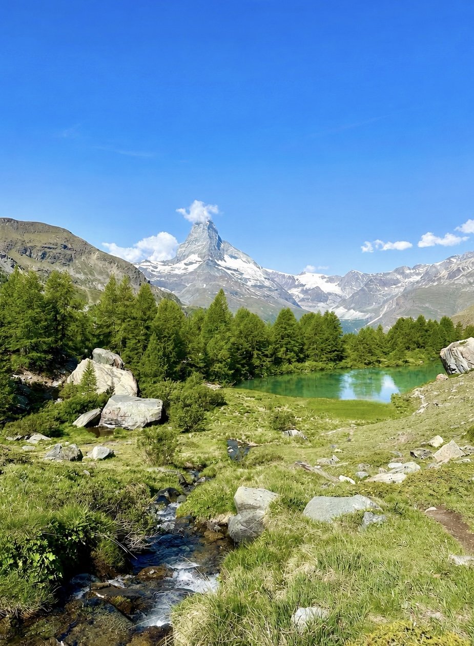 A scenic mountain landscape featuring a snow-capped peak, green pine trees, a small turquoise lake, and a flowing stream in a lush meadow under a bright blue sky with some clouds. Five lakes hike, Zermatt, Matterhorn.