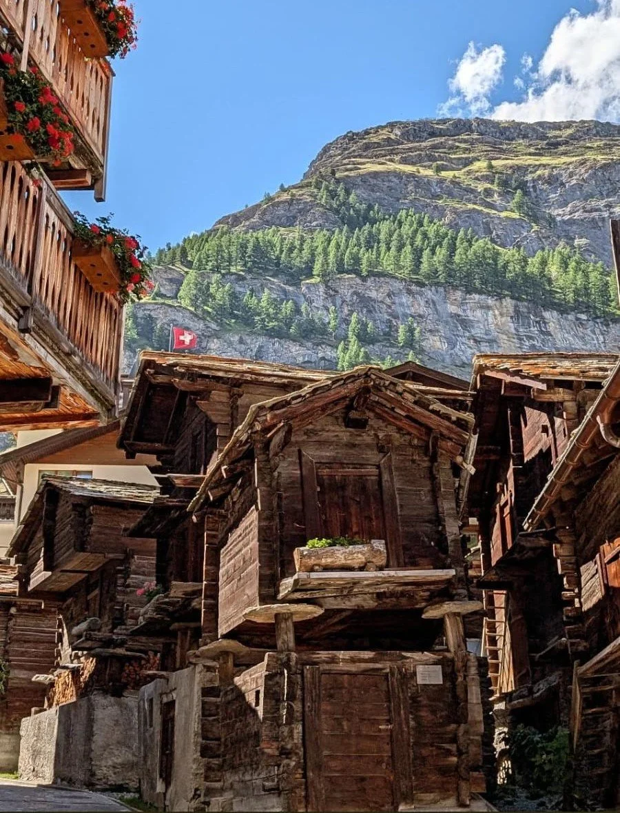 Traditional Swiss chalets made of wood in a mountain village with a large mountain and forest in the background, and a Swiss flag on a flagpole. Zermatt.