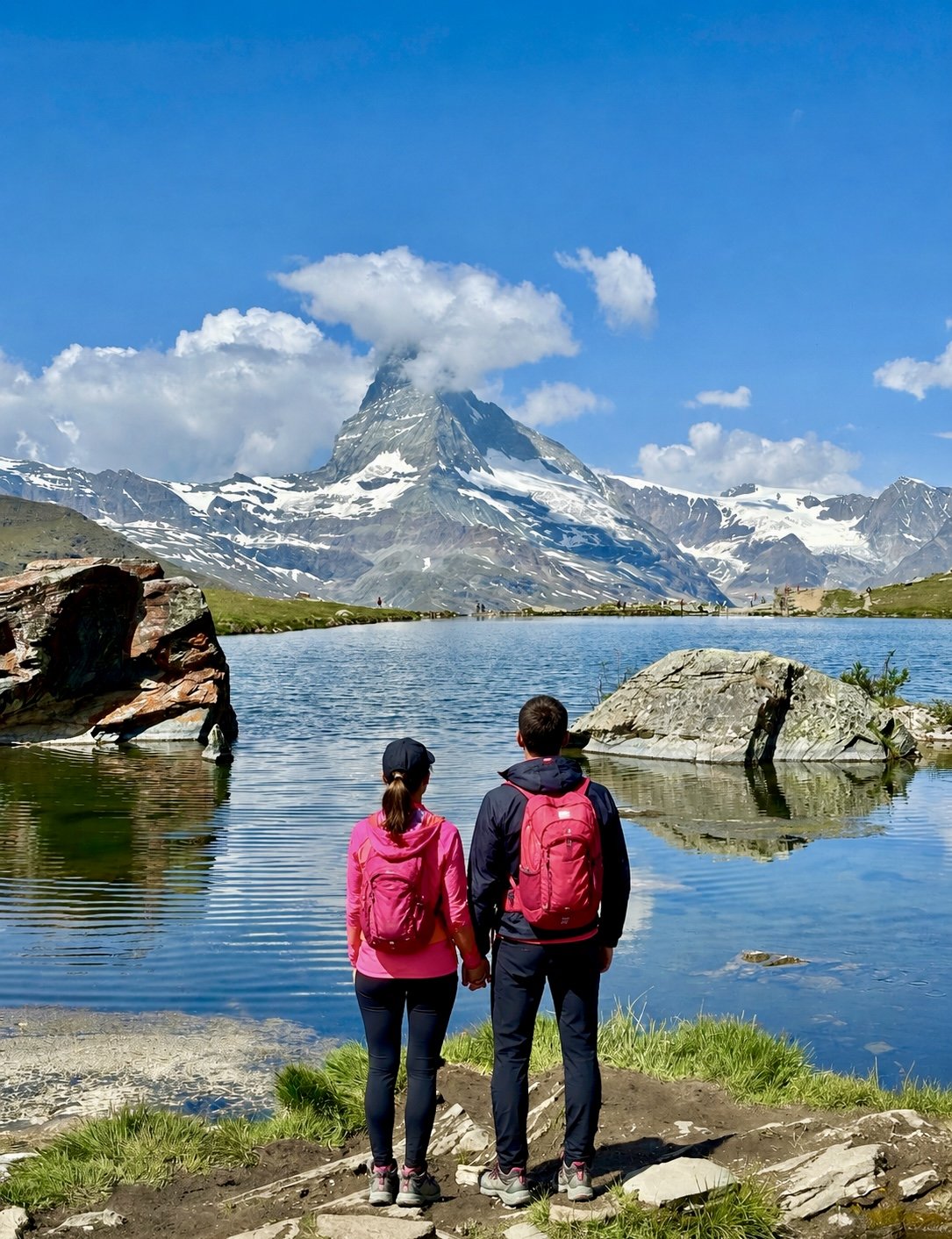 A couple with backpacks standing by a lake and looking at a snow-capped mountain in the distance on a bright, partly cloudy day. Five lakes hike, Zermatt, Matterhorn.