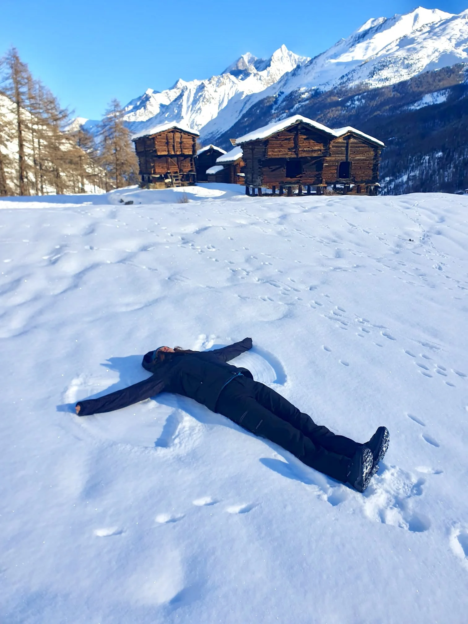 A person lying on snow in a winter landscape with snow-covered mountains and trees in the background. Zmutt, Zermatt.