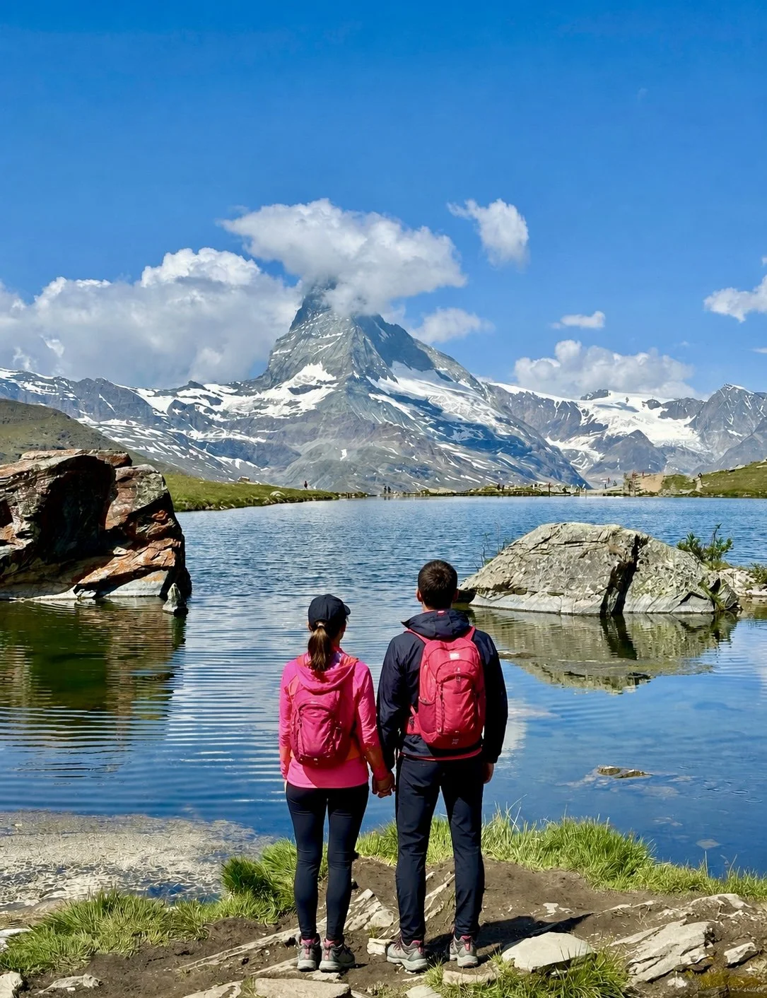 A man and woman with backpacks stand hand-in-hand by a lake, on a Five- lakes hike in Zermatt, overlooking Matterhorn with snow patches and clouds at the summit, under a blue sky.