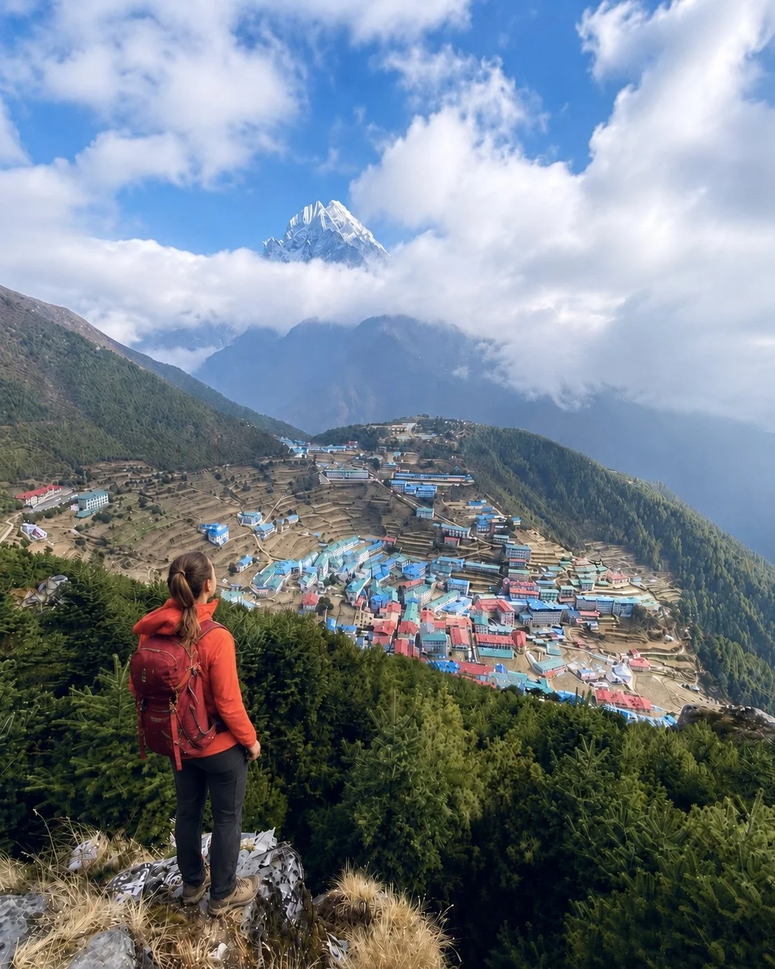 A woman with a red jacket and backpack standing on a hill overlooking a colorful village on a terraced mountainside, with snow-capped peaks and clouds in the background. Everest Base Camp trek.