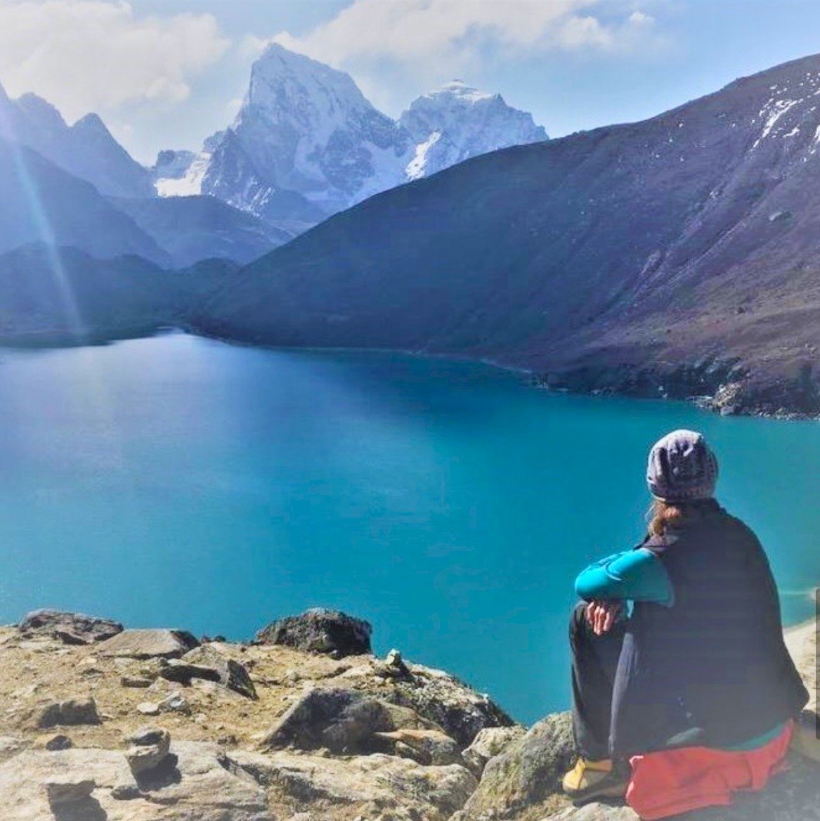 Person sitting on rocks, looking at a large blue lake surrounded by mountains, including snow-capped peaks in the distance. Everest Base Camp trek, Gokyo lake.