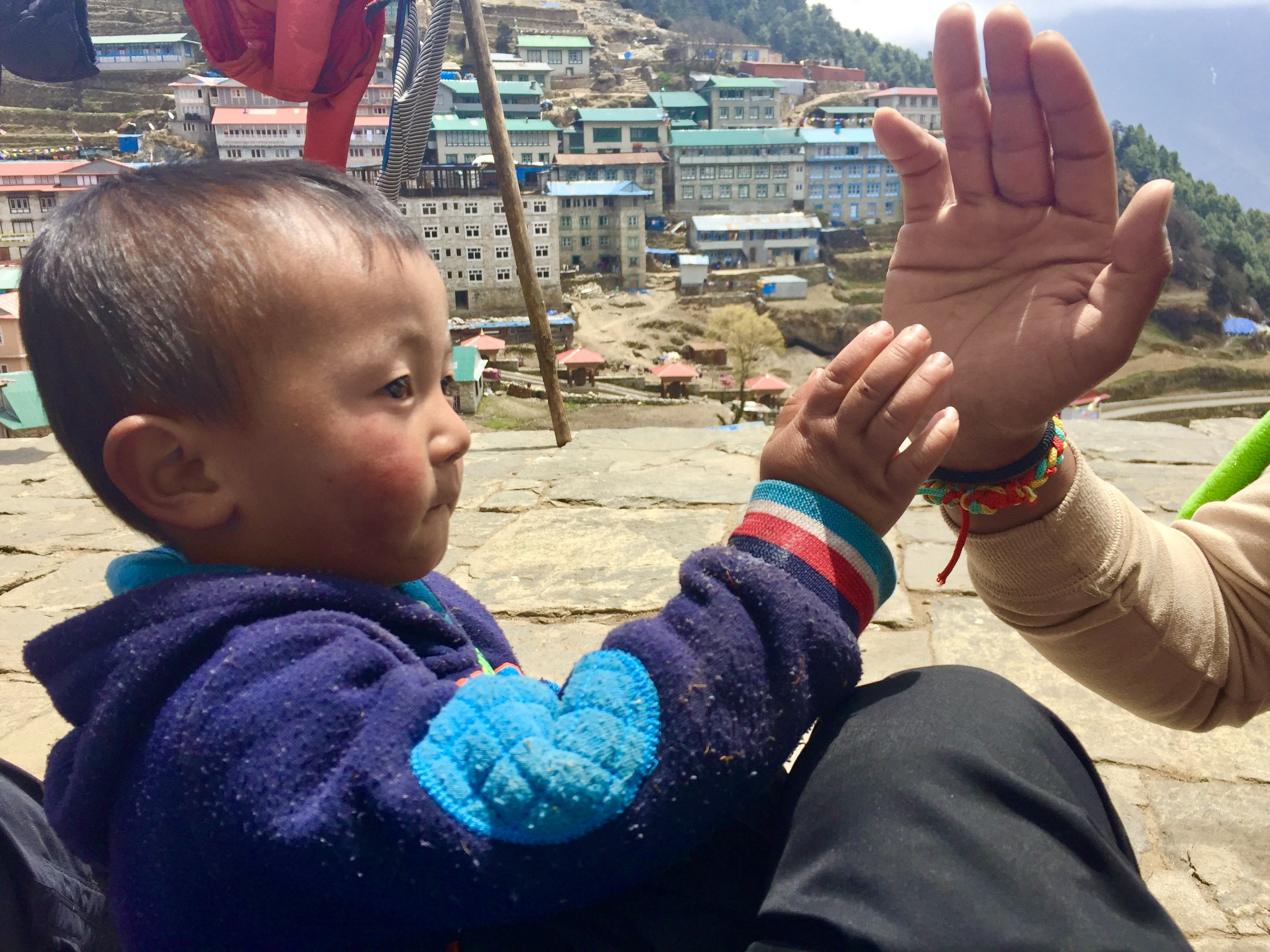 A young child with dark hair and wearing a blue and purple fleece jacket sitting outdoors. An adult with a tan sweater and colorful wristbands is extending their hand towards the child. In the background, there are colorful buildings on a hillside, suggesting a mountainous or hilly region.