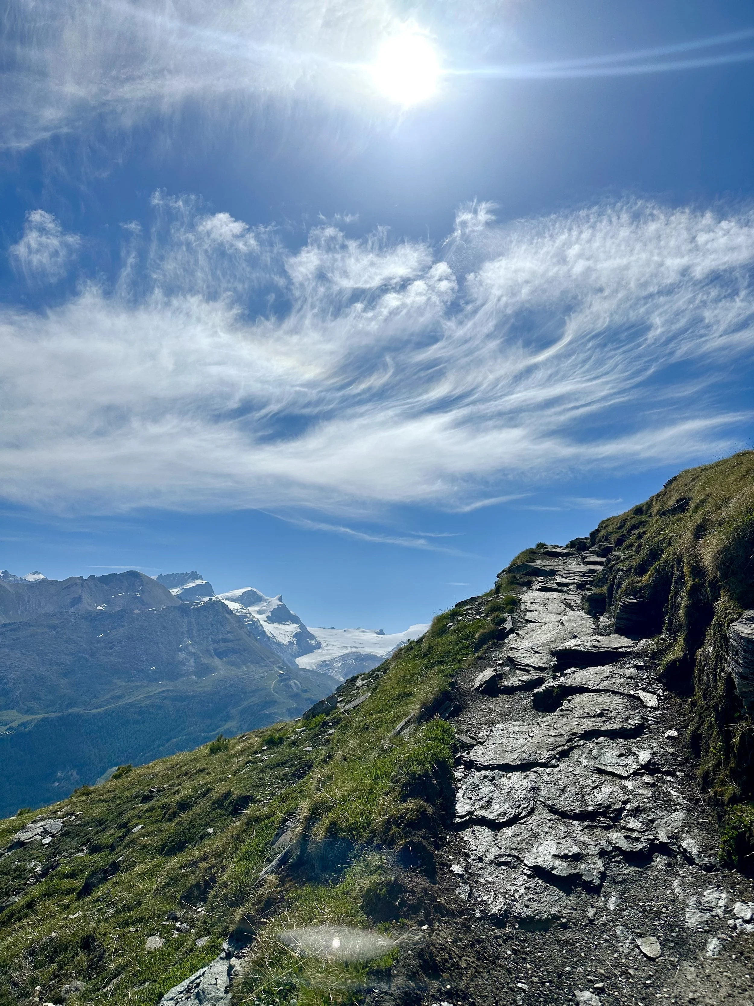 A mountain trail on a grassy slope with rocky steps, with snow-capped mountains in the distance and a bright sun in the clear blue sky. On a High-trail or Höhenweg in Zermatt.