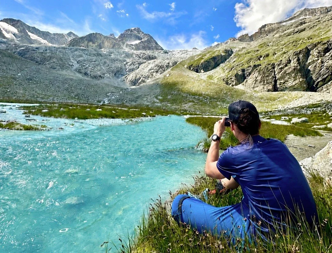 A person sitting by a turquoise mountain lake taking a photo of the scenery with mountains and a partly cloudy sky in the background. Five lakes hike, Zermatt.