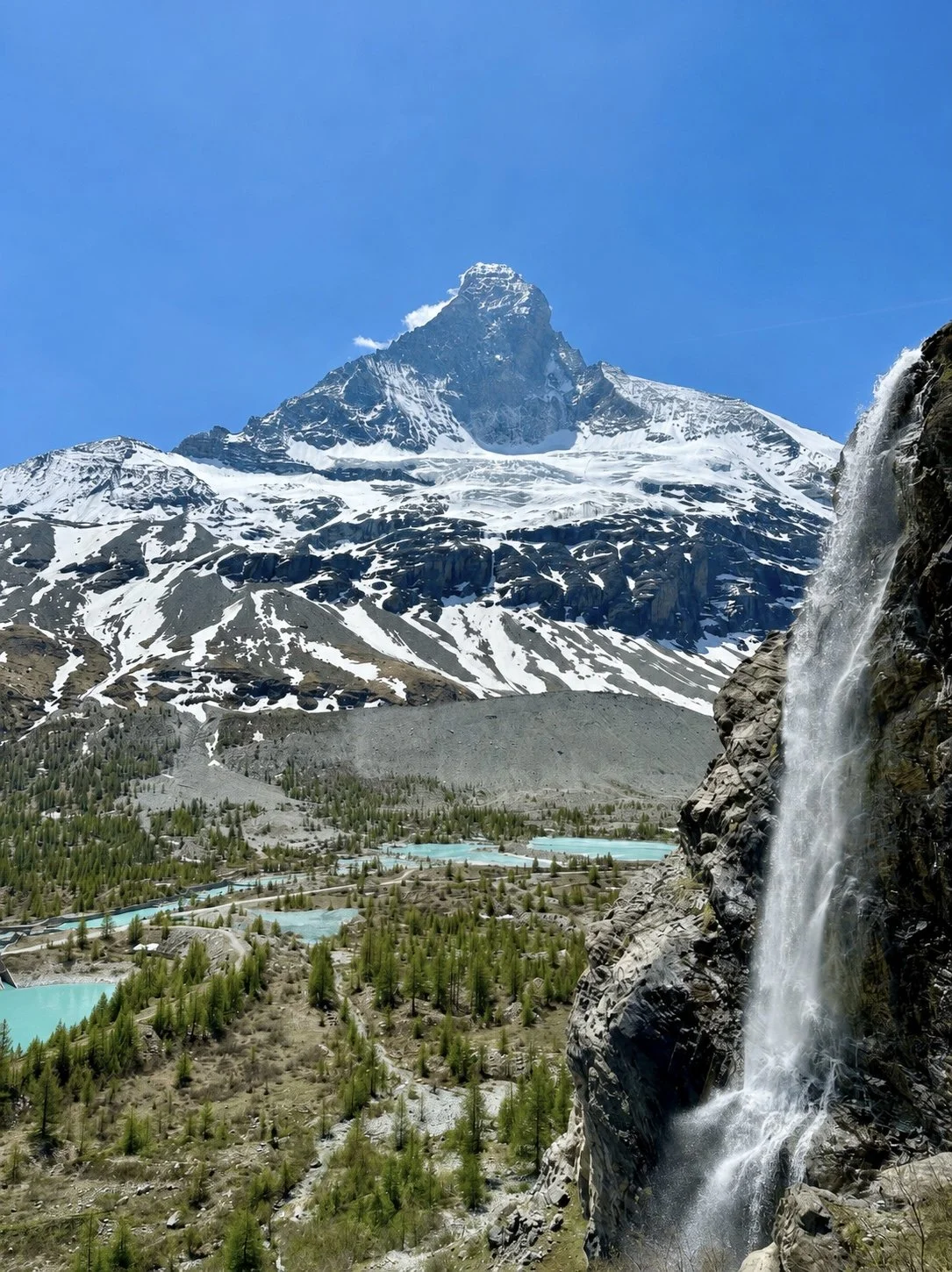 Snow-capped mountain with sharp peak, waterfall on rocky cliff, and lakes in the green valley below under a clear blue sky. North face Matterhorn, Zermatt.