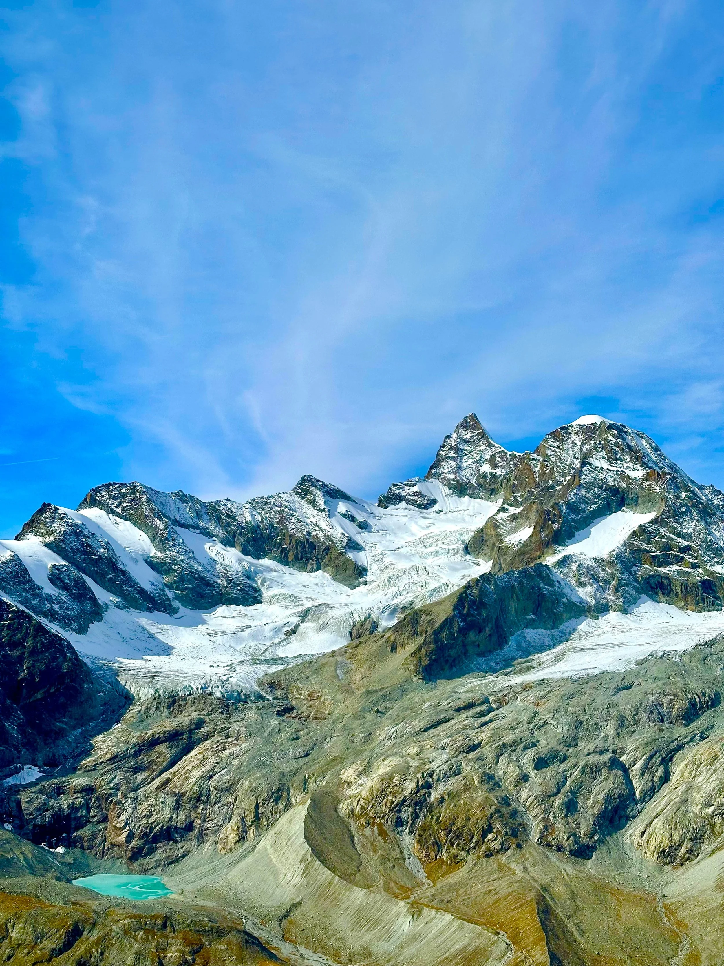 Snow-capped mountain peaks under a blue sky with wispy clouds, rugged terrain, and a small turquoise lake at the base. High trail hike, Höhenweg, Zermatt.