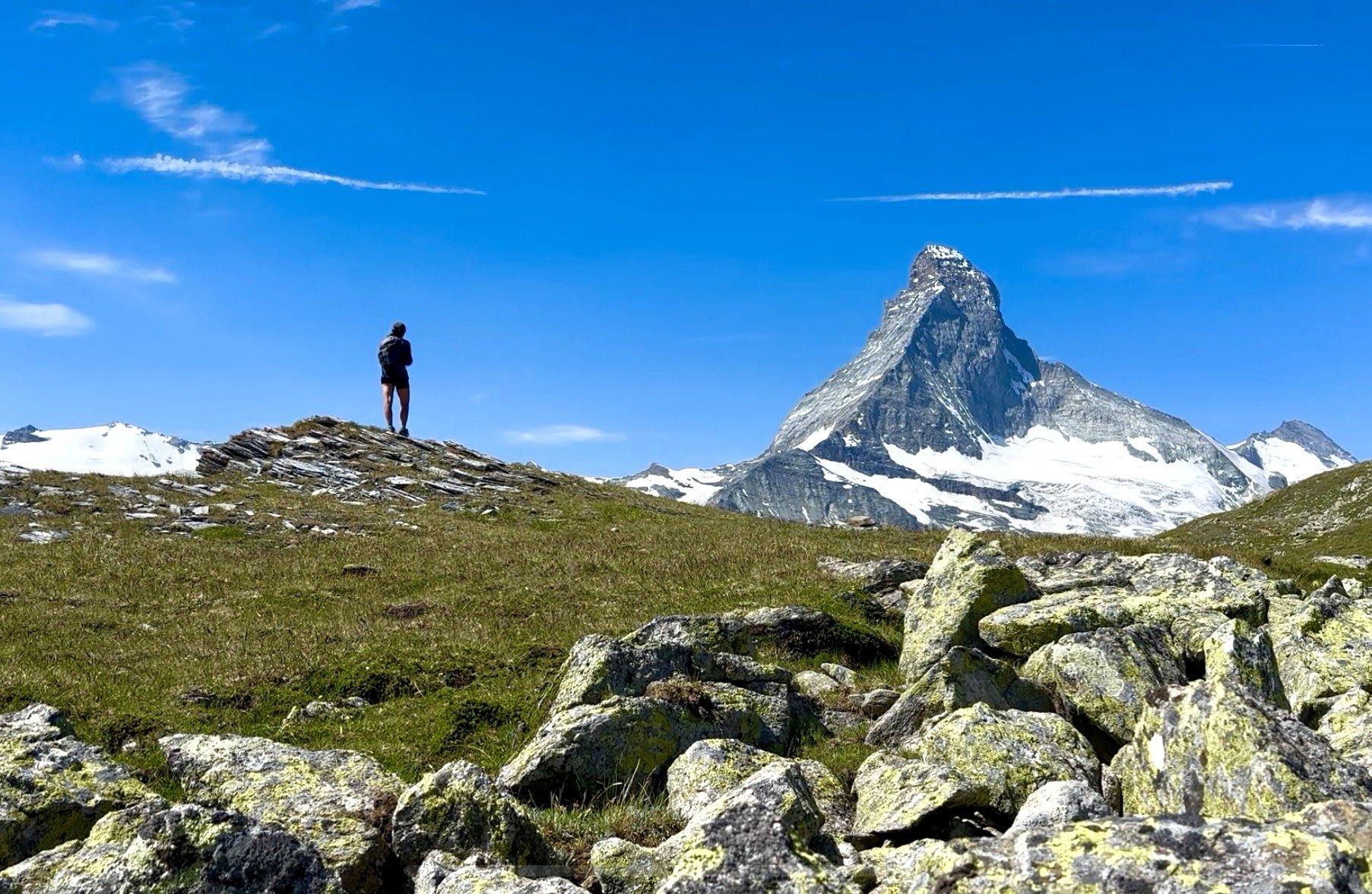 A hiker standing on a grassy and rocky landscape with snow-capped mountains and a blue sky with a few scattered clouds in the background. High trail hike, Höhenweg, Zermatt.