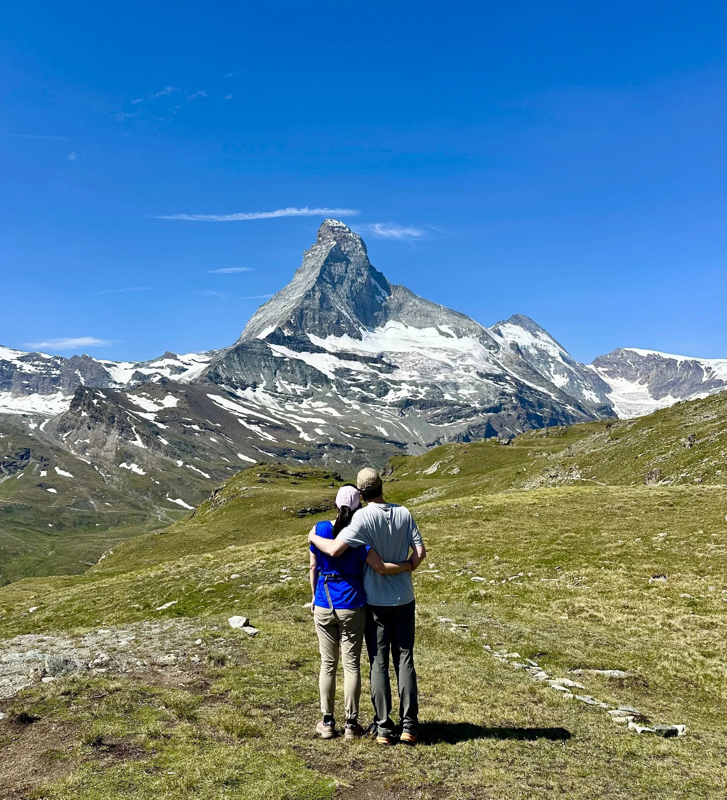 A couple stands close together on a mountain trail, surrounded by mountainous terrain and snow patches, with a prominent mountain peak in the background under a bright blue sky. High trail hike, Höhenweg, Zermatt.