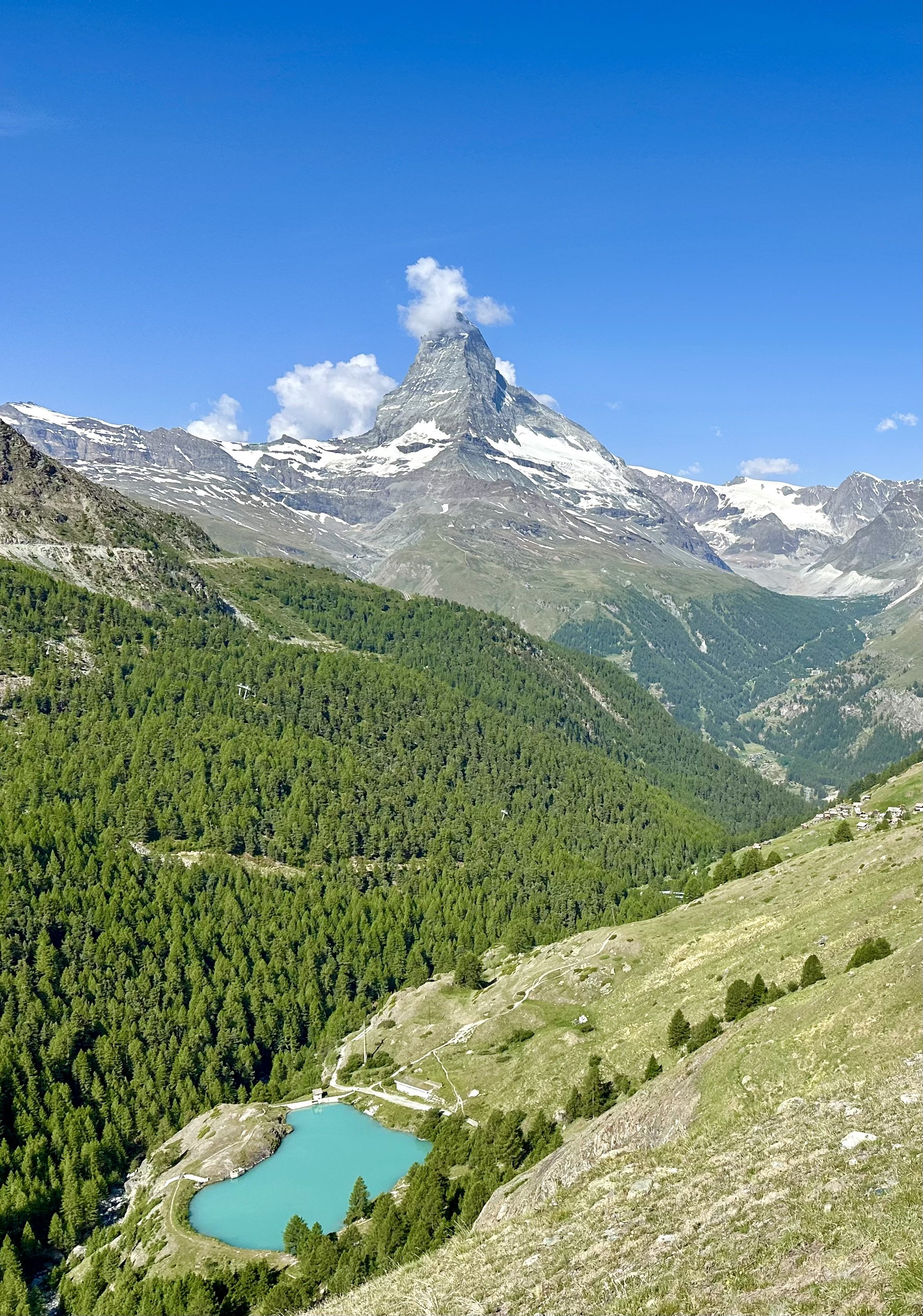 Snow-capped Matterhorn, surrounded by other mountainous terrain, under a clear blue sky. A forested valley with a turquoise lake and a small building are visible in the foreground. On a Five-lakes hike, Zermatt.