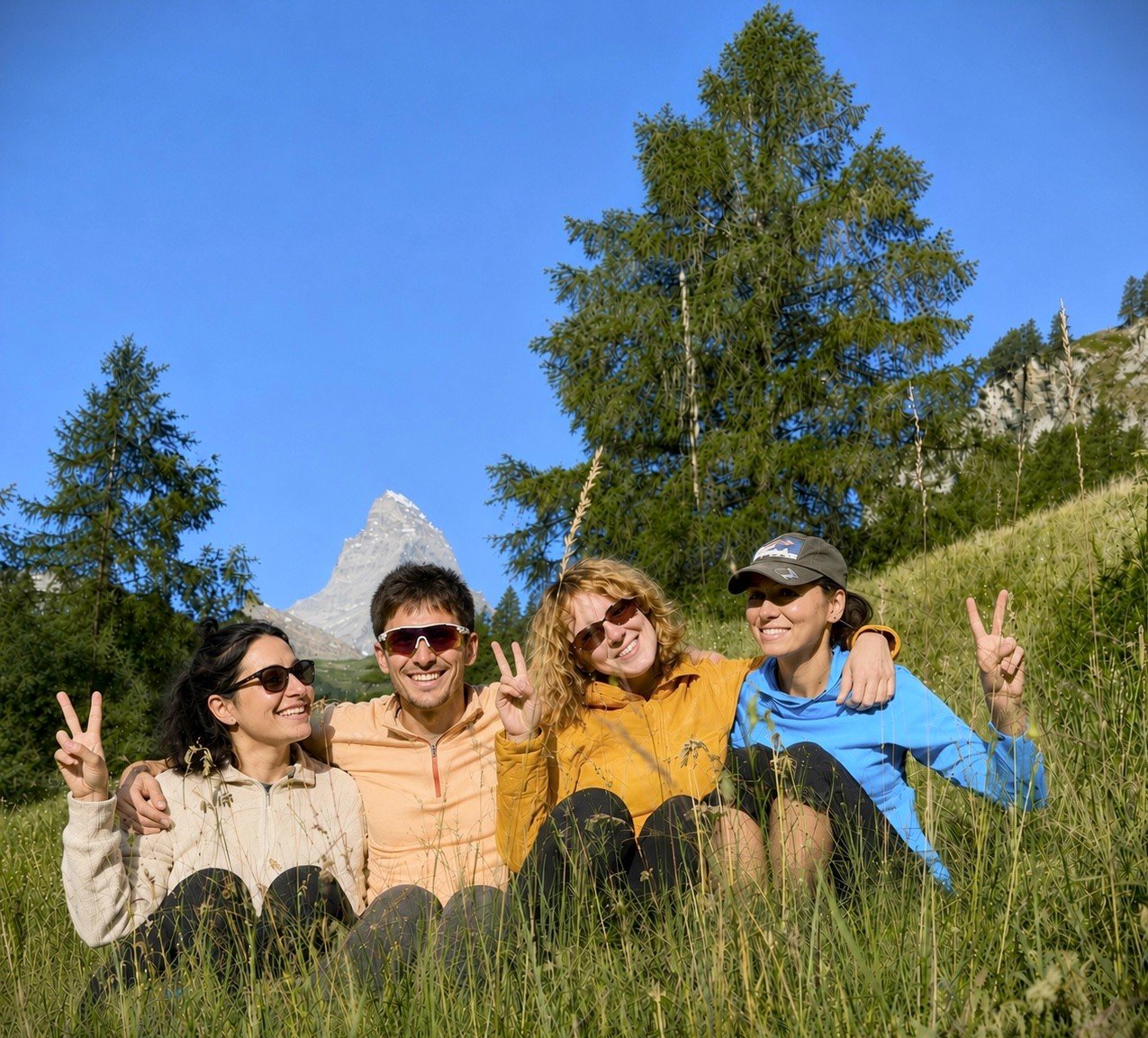 Four friends sitting on grass in a mountain meadow, smiling and making peace signs, with trees and a mountain peak in the background.