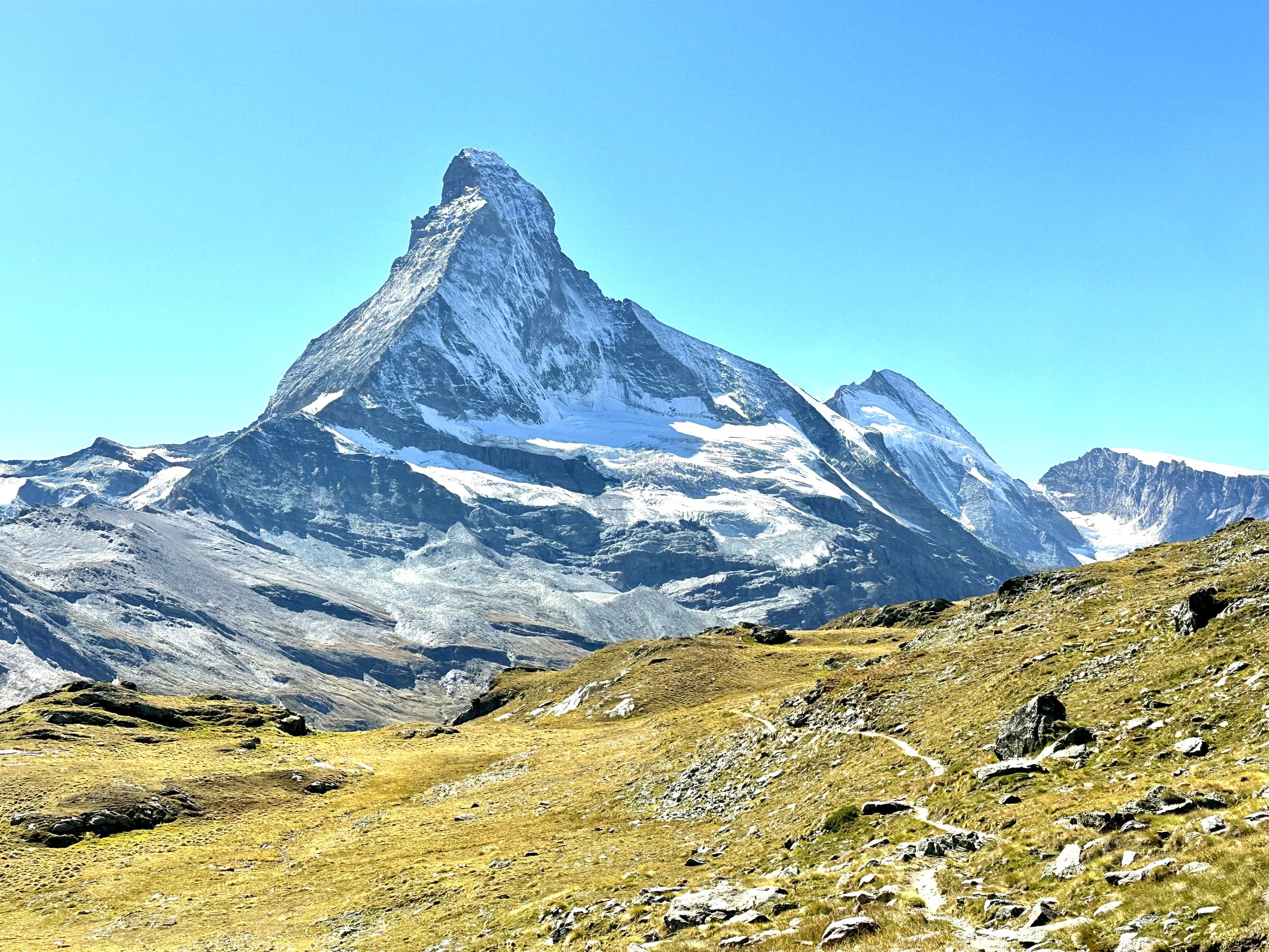 Snow-covered mountain peak with rocky slopes and grassland foreground under clear blue sky. Matterhorn, Zermatt, High trail hike.