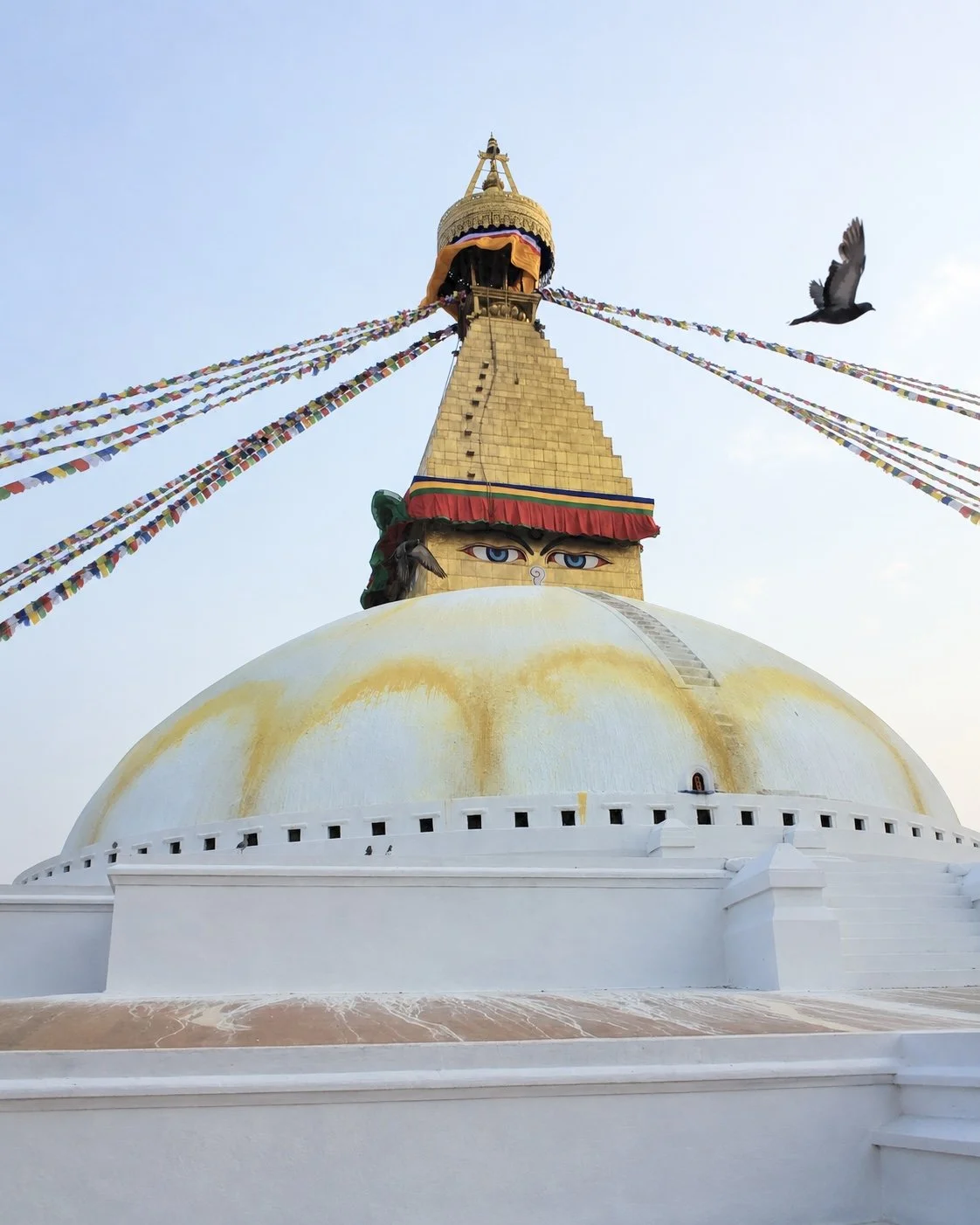 Low-angle view of a large white stupa with a golden spire topped with a dome decorated with colorful prayer flags, against a light blue sky. The stupa has painted eyes and a ladder leading to the top, with a bird flying nearby. Everest Base Camp trek