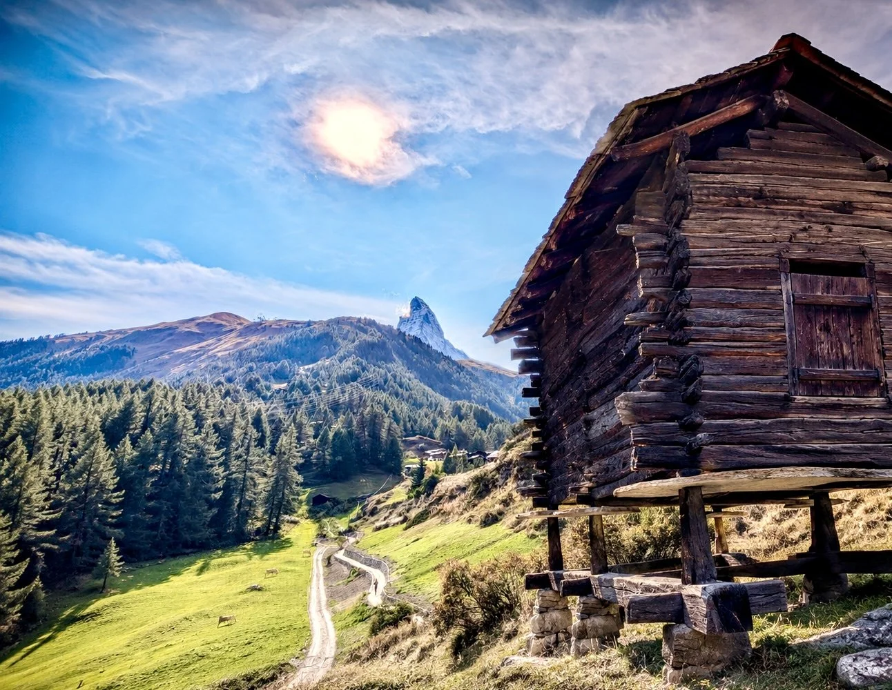 A rustic wooden cabin on stilts overlooking grassy rolling hills and a forest, with mountains and a bright blue sky with clouds and the sun in the background. Zmutt, Culturweg, Zermatt.