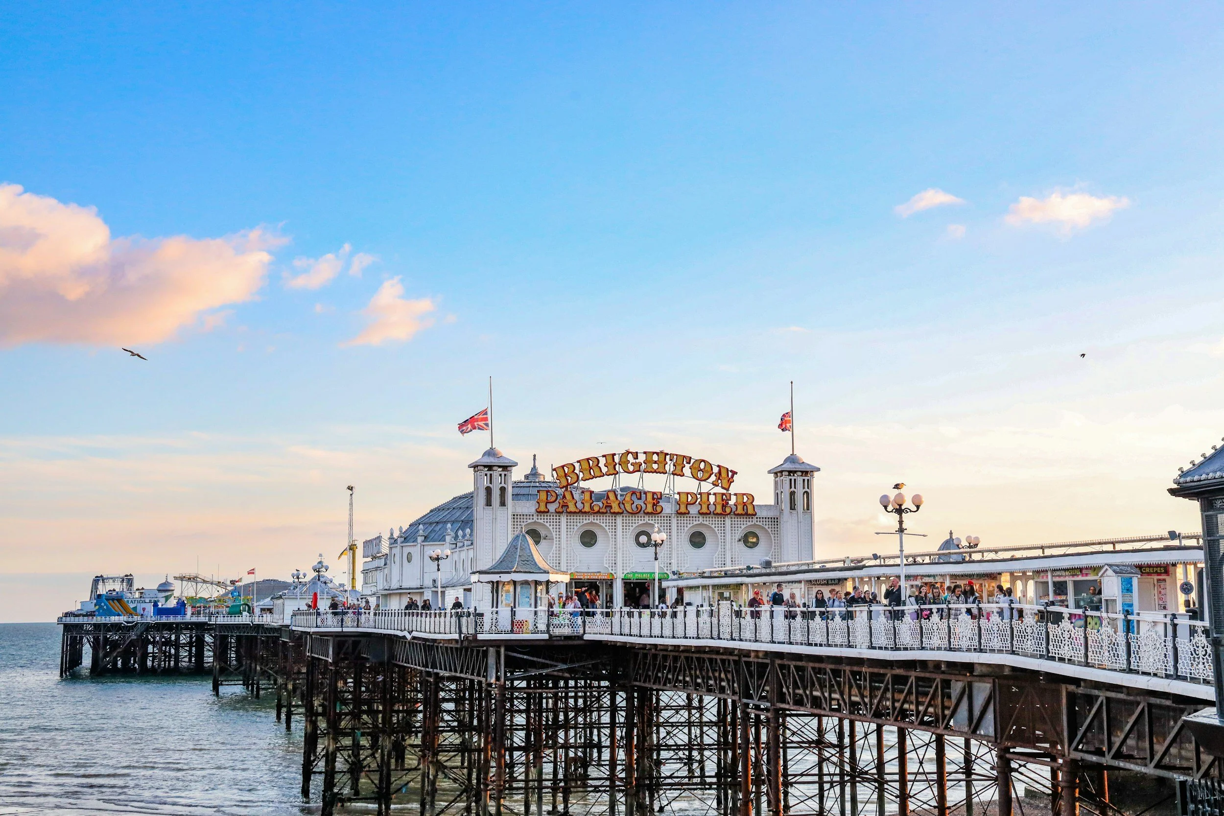 Brighton Palace Pier with its white Victorian-style building and signage, flags, and amusement rides on the pier, with a clear blue sky and a few clouds in the background.