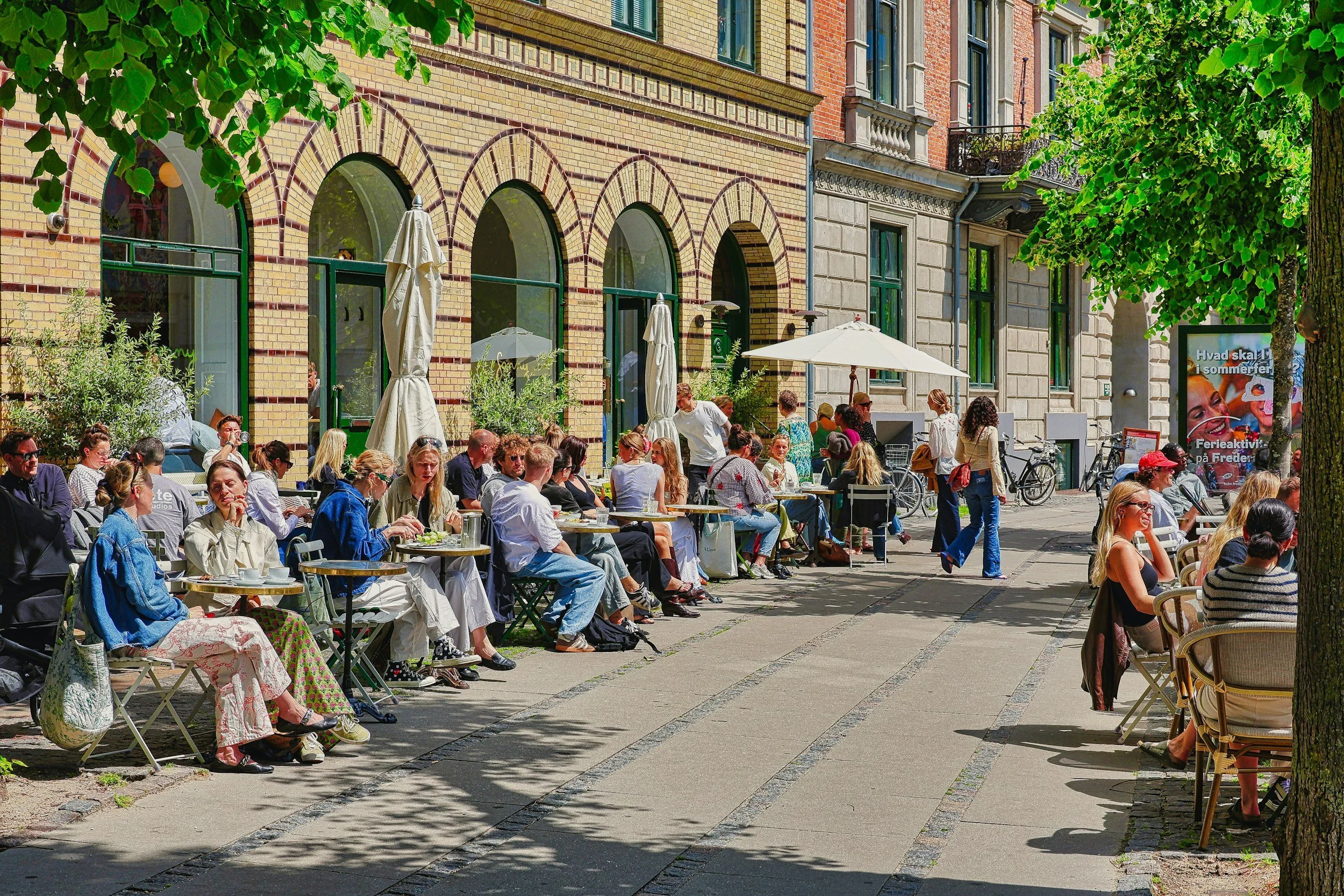 A crowded outdoor cafe with people seated at tables on a sunny day, surrounded by buildings with brick and stone facades, and trees providing shade.