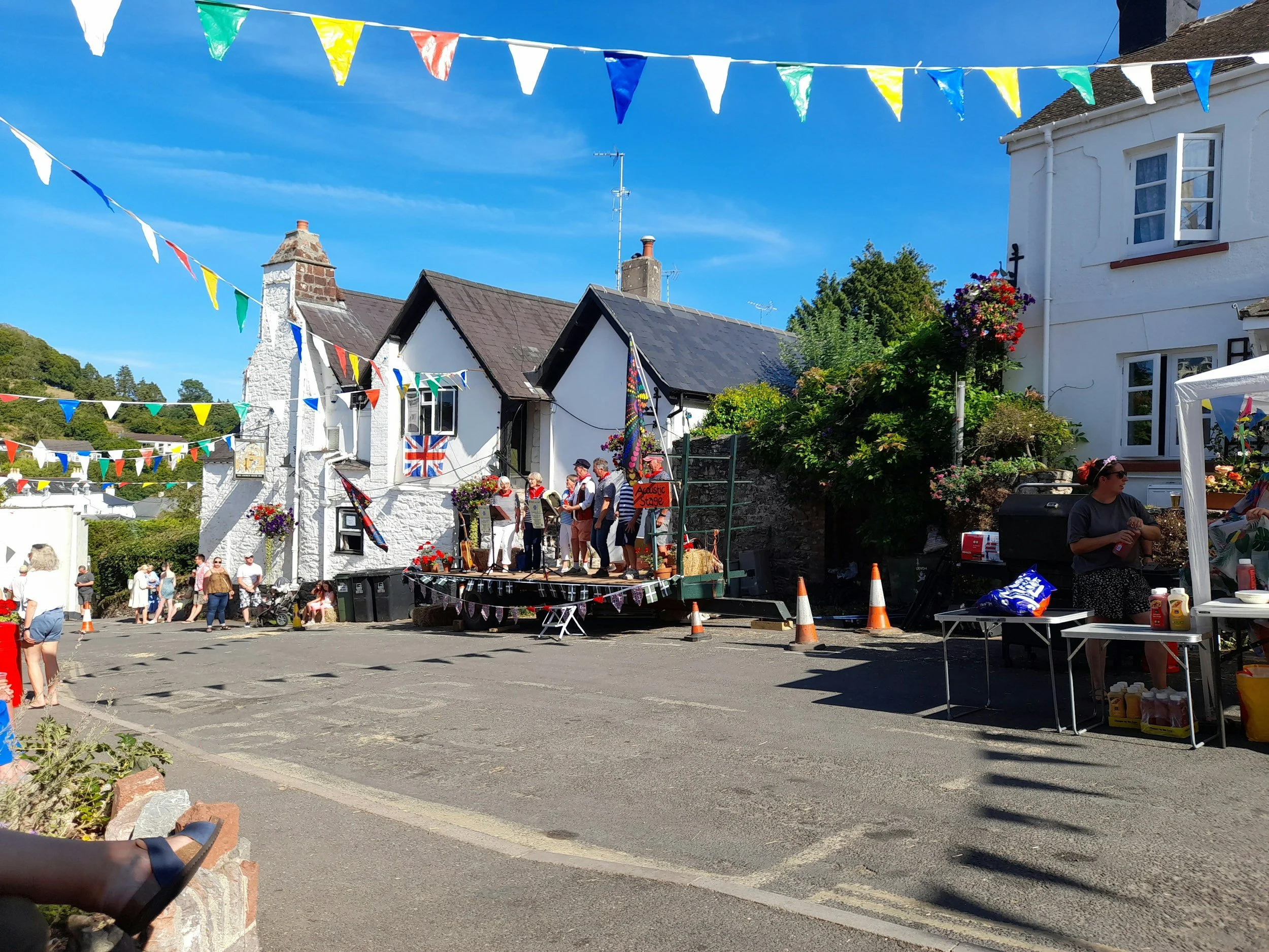 A street scene with bunting flags hanging above and a small stage set up with people gathered, possibly for a community event or festival, in front of white buildings with a blue sky overhead.