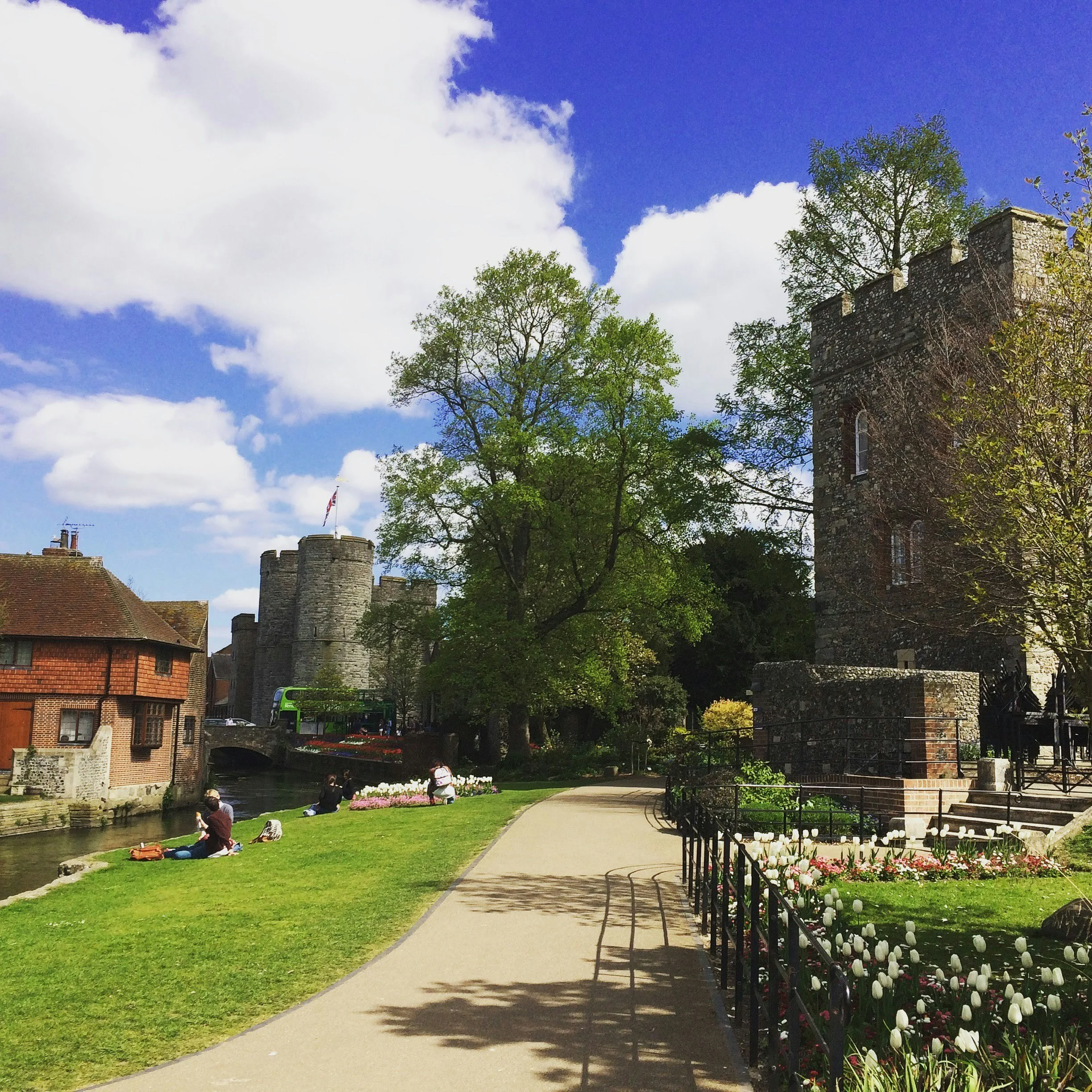 People sitting on the grass near a river, with historic stone towers and buildings in the background under a partly cloudy sky.