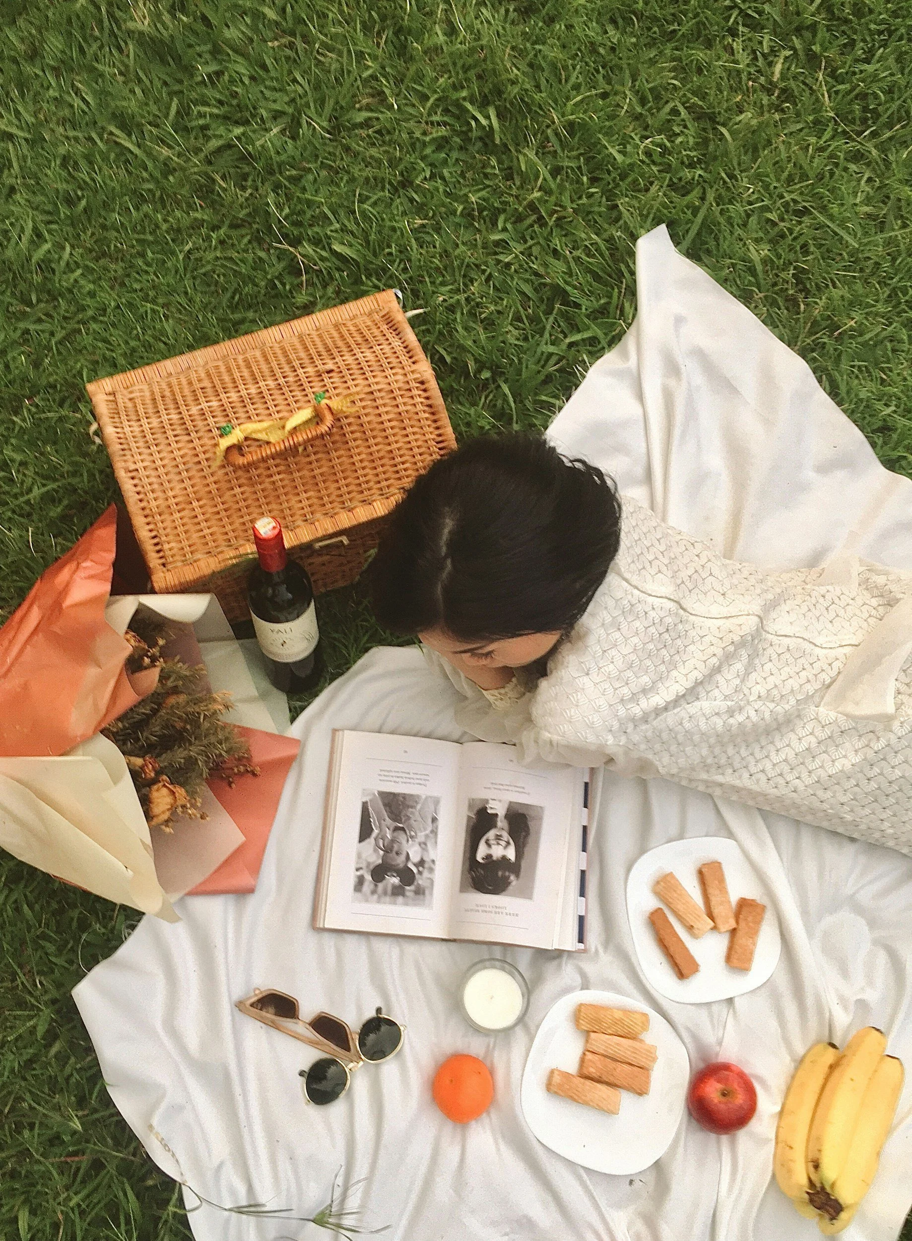 A picnic setup on a white cloth on grass, featuring a wine bottle, a book with black-and-white photographs, plates with snacks including crackers, an apple, bananas, candles, sunglasses, and floral arrangements.