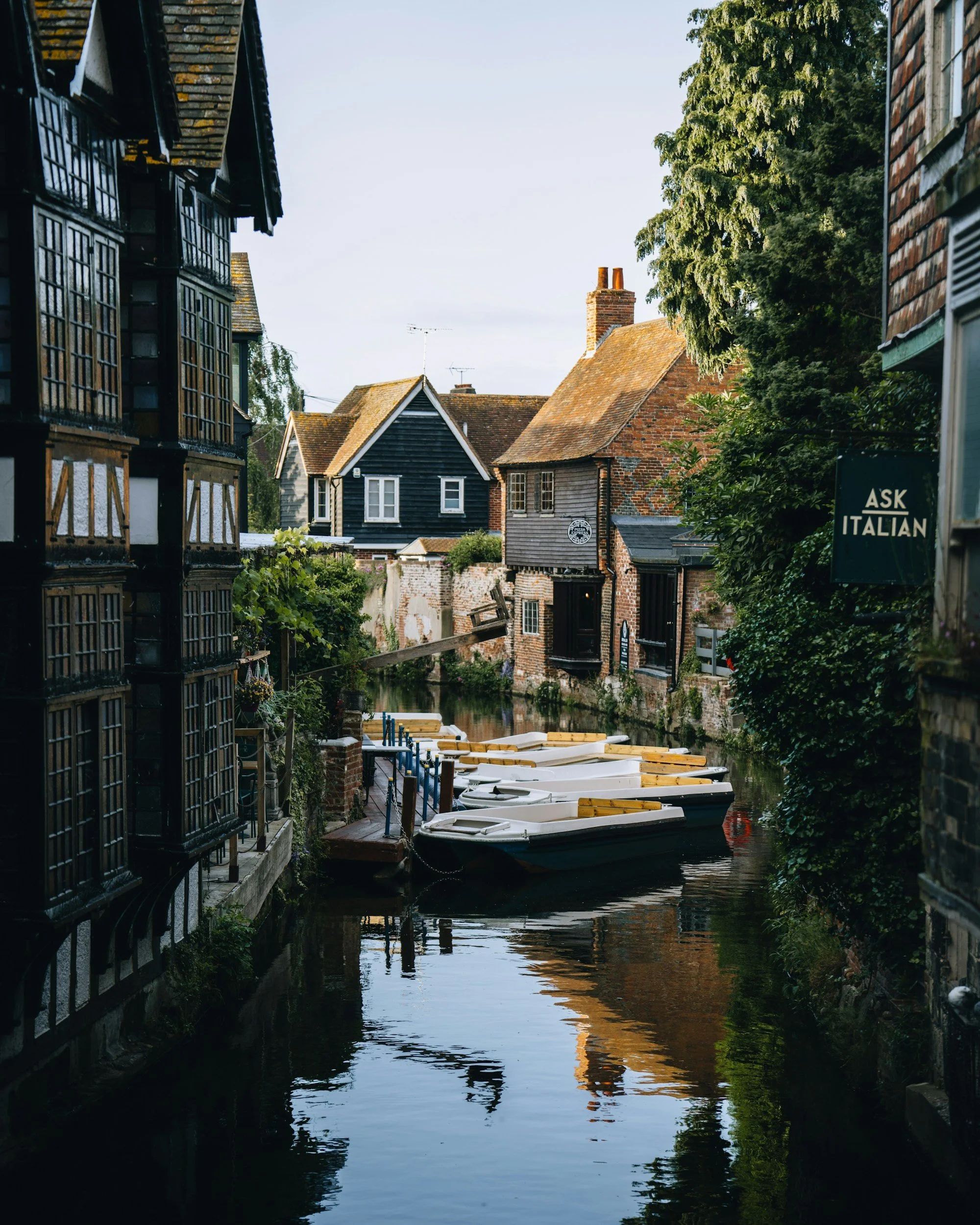 A narrow canal lined with old buildings, some with timber framing and brick walls, and small boats docked along the edge, with trees on the right side and a sign that says 'Ask Italian' on the right.