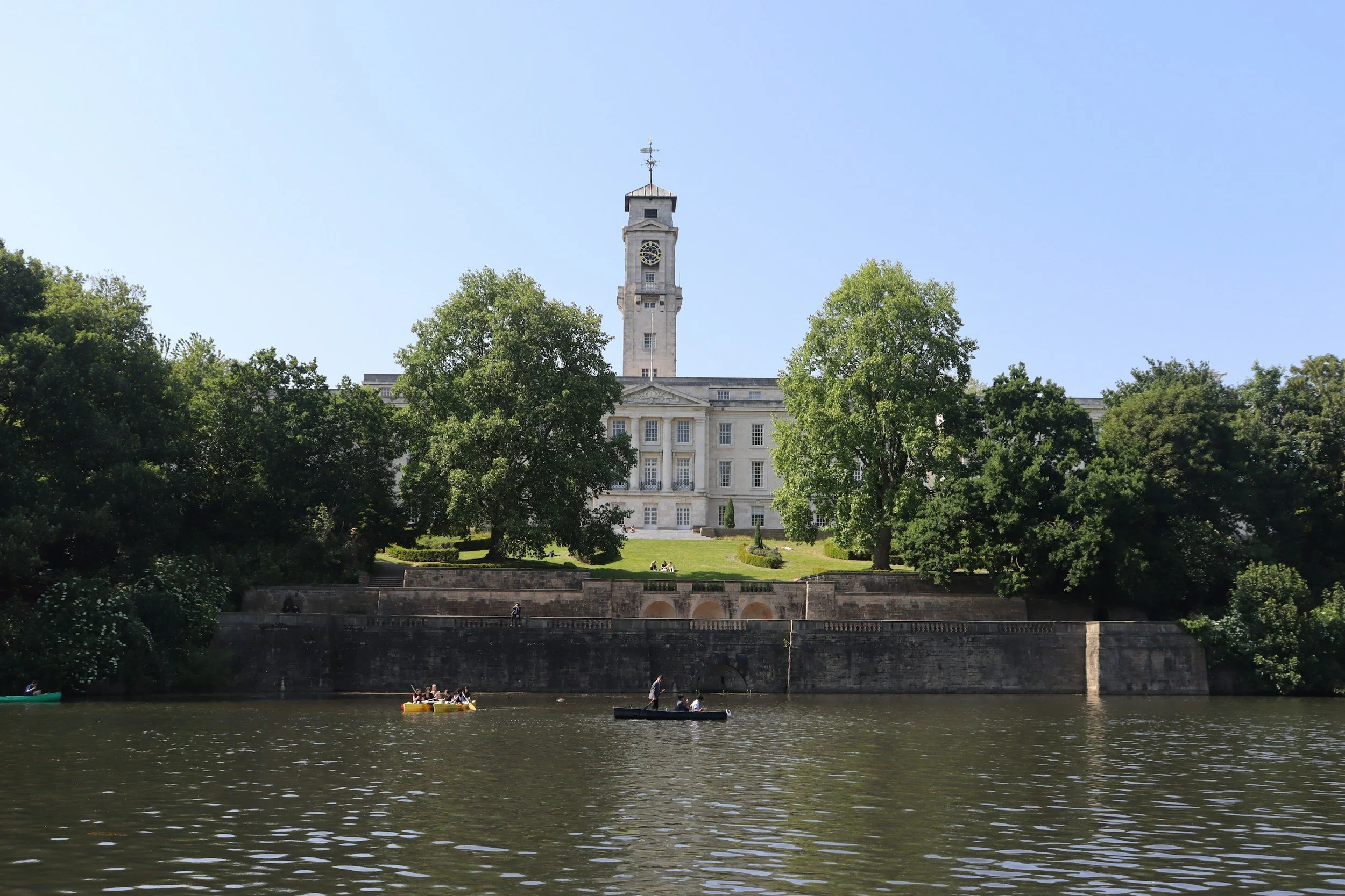 A historic building with a clock tower surrounded by green trees, viewed across a river with people kayaking and canoeing.