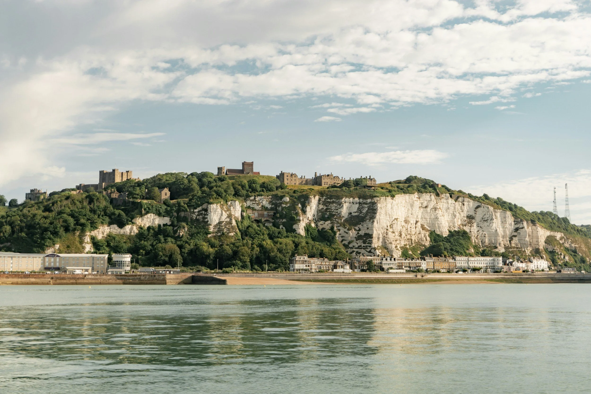 A castle and several buildings situated on a steep hill with greenery, overlooking a body of water under a partly cloudy sky.