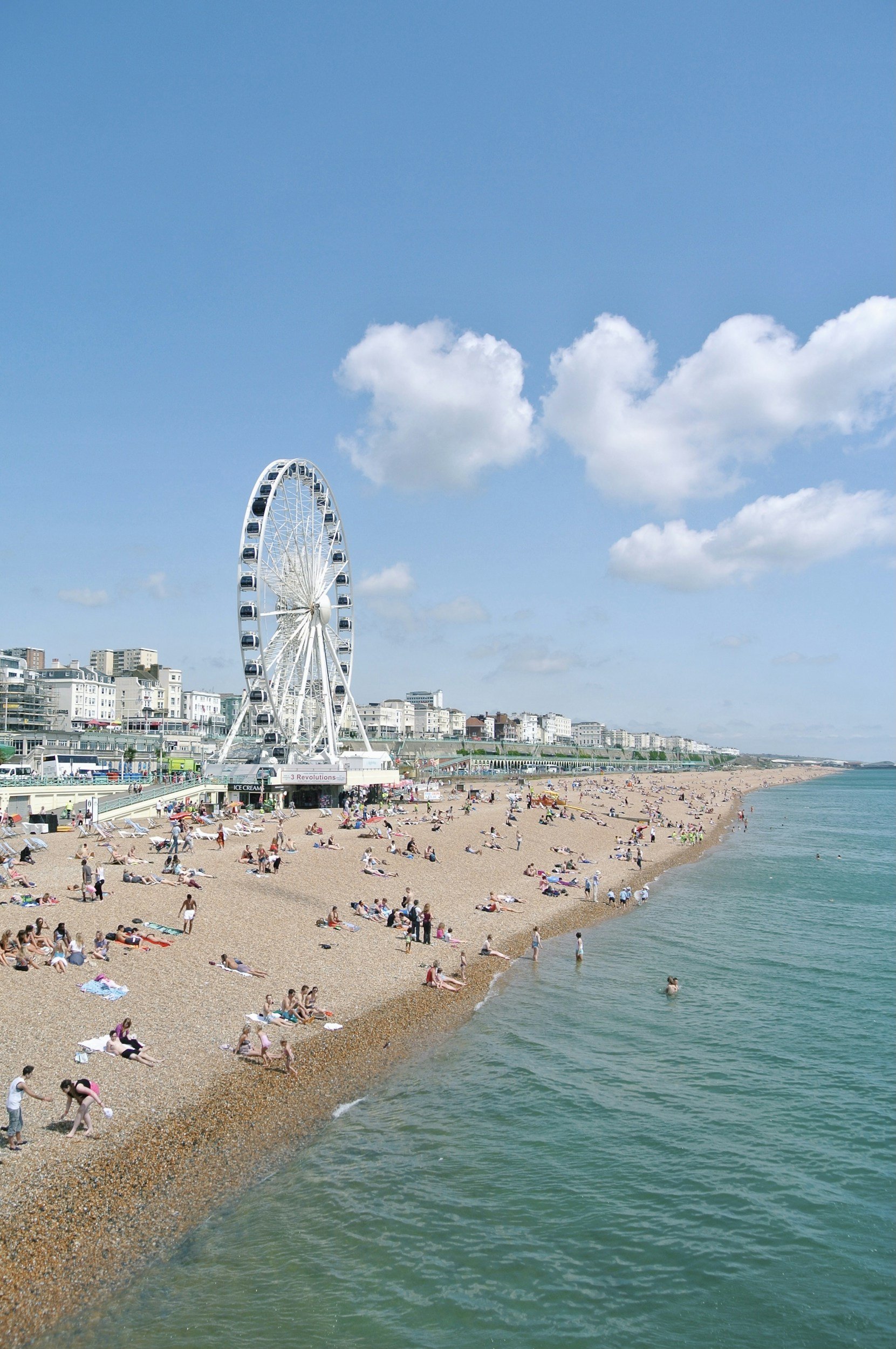 A crowded beach with people lounging on the sand, near the water, and walking along the shore. A large Ferris wheel is visible in the background against a blue sky with scattered clouds. City buildings are seen further inland.