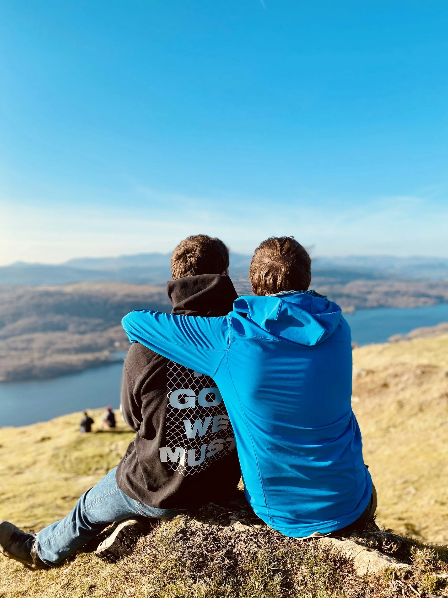 Two men sitting on a hilltop overlooking a lake and mountains on a clear day, one with arm around the other.