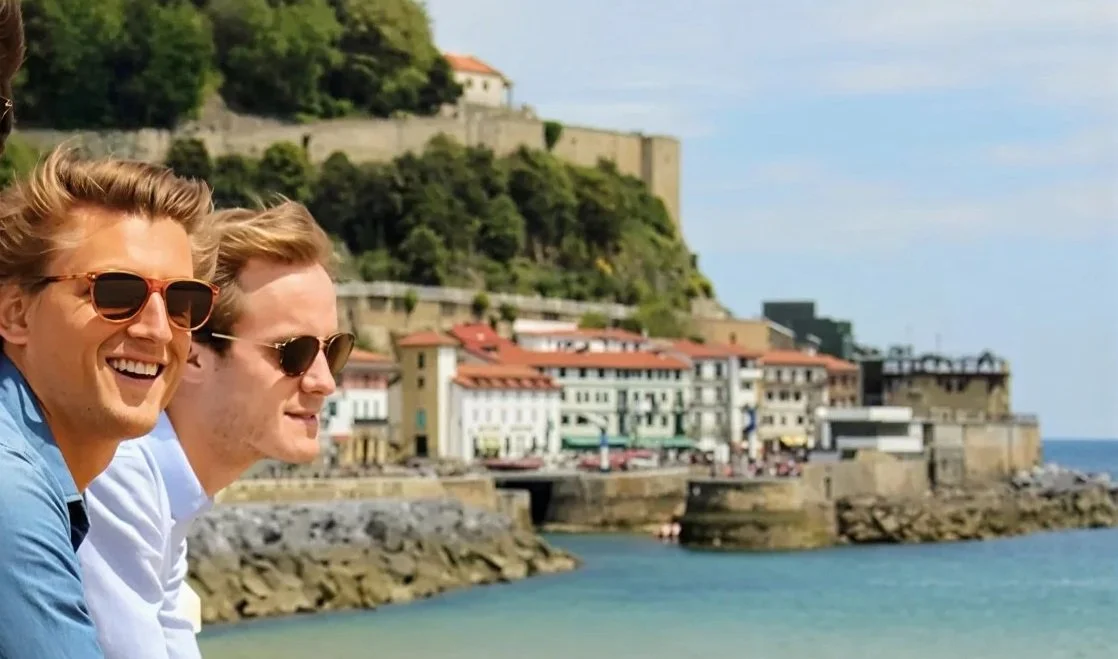 Two young men wearing sunglasses smiling by the seaside with a historic coastal town and castle in the background.