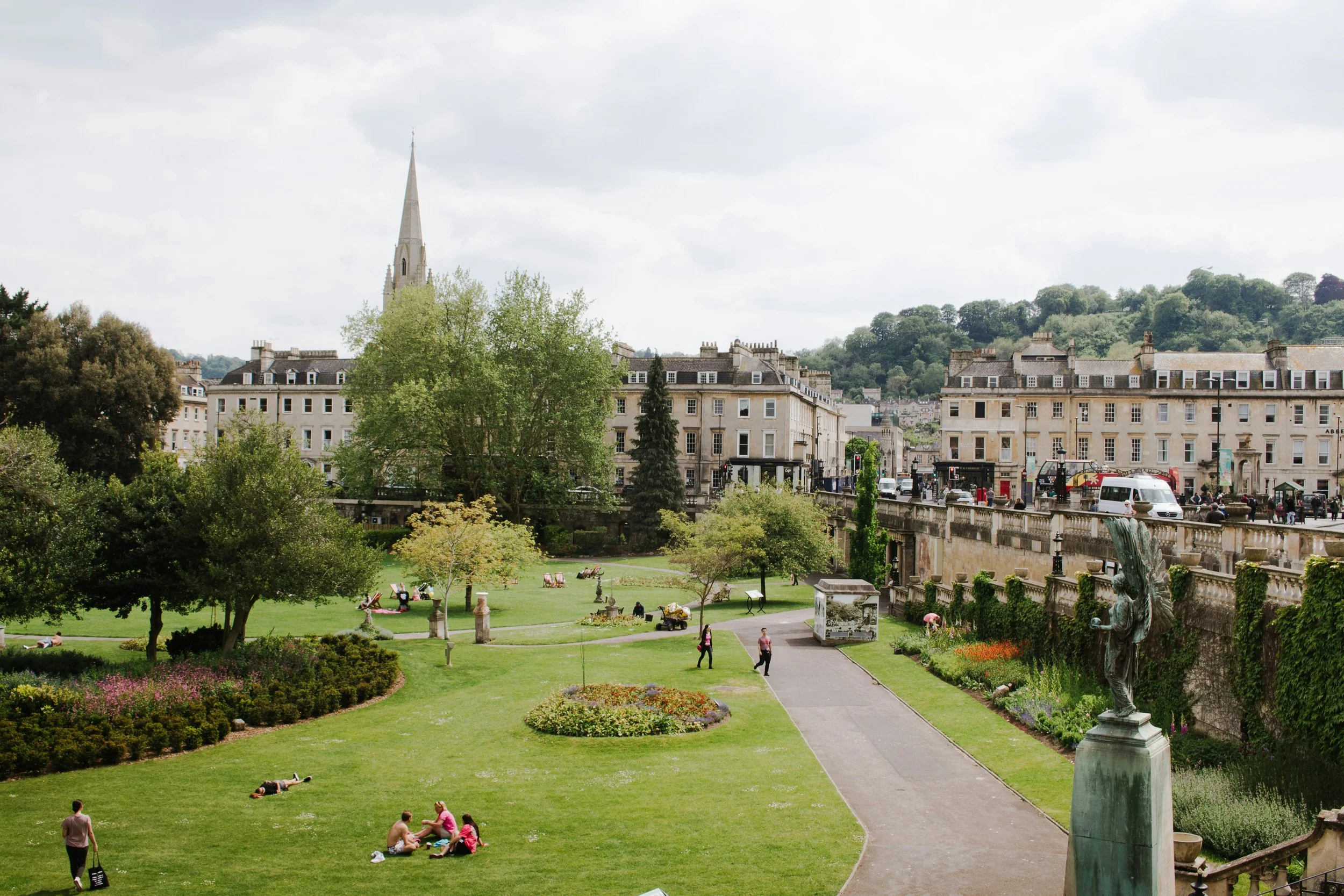 A park with green grass, trees, walking paths, and benches, with people lounging and walking. There is a row of classic buildings and a church steeple in the background, with hills and cloudy sky above.