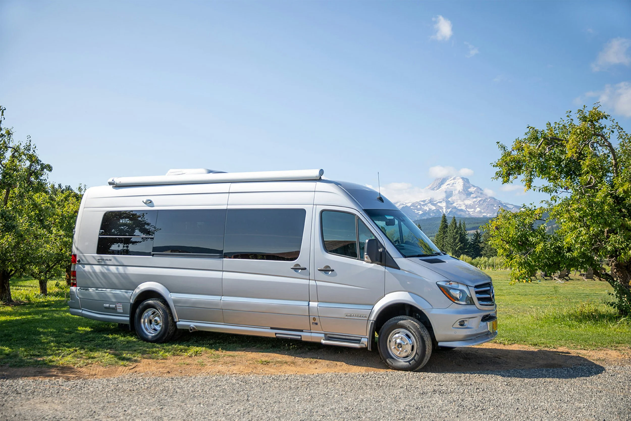 Silver camper van parked on a grassy area with trees, mountains, and a blue sky in the background.