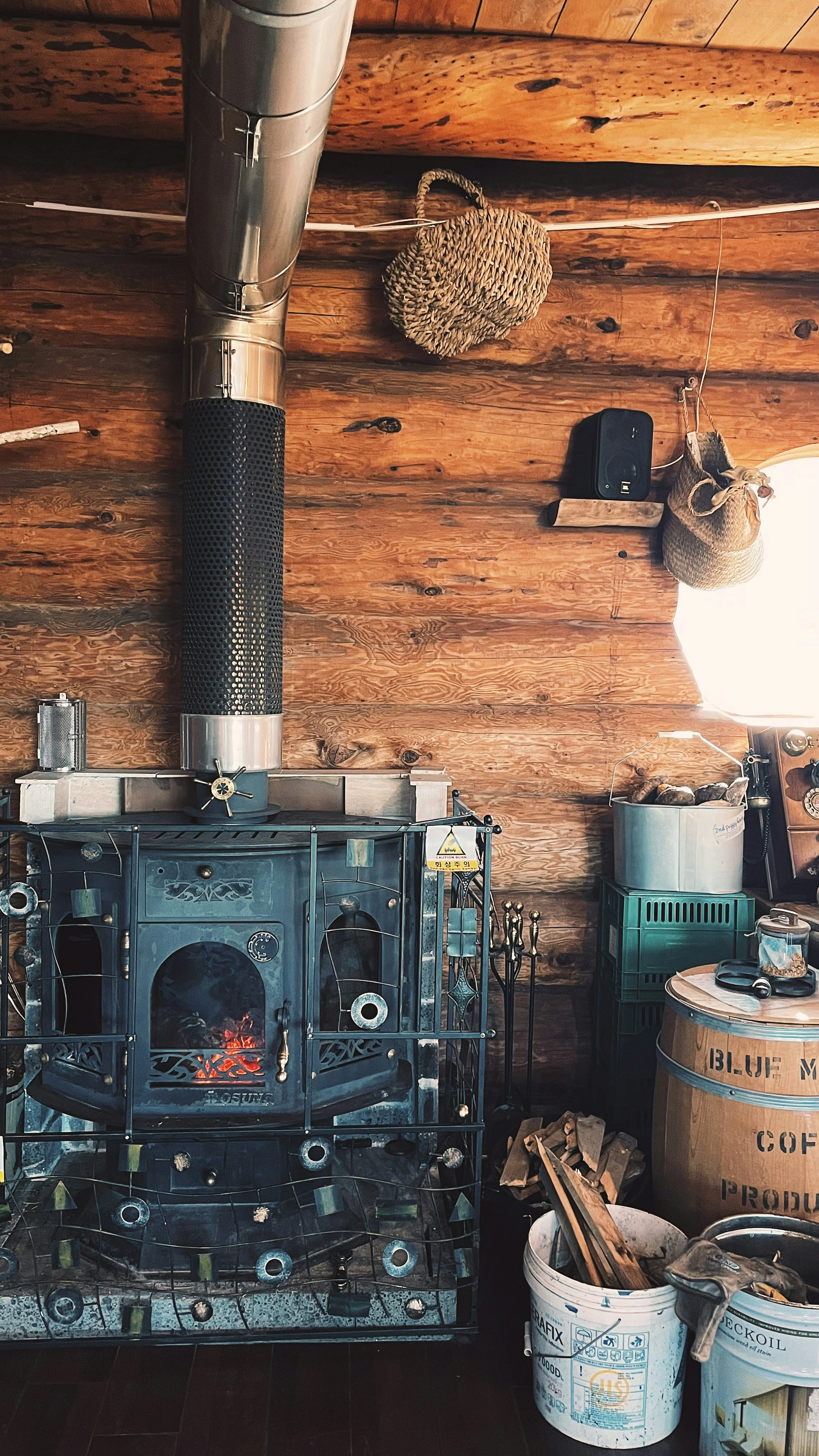 A cozy rustic room with wooden walls and ceiling. A black metal stove with a fire inside in the foreground. Above the stove, a silver chimney pipe extends up to the ceiling. There are two woven baskets hanging on the wall, and a black speaker mounted on the wall. To the right, a wooden table, a barrel labeled 'Blue Moon Coffee', and several buckets containing firewood and tools.