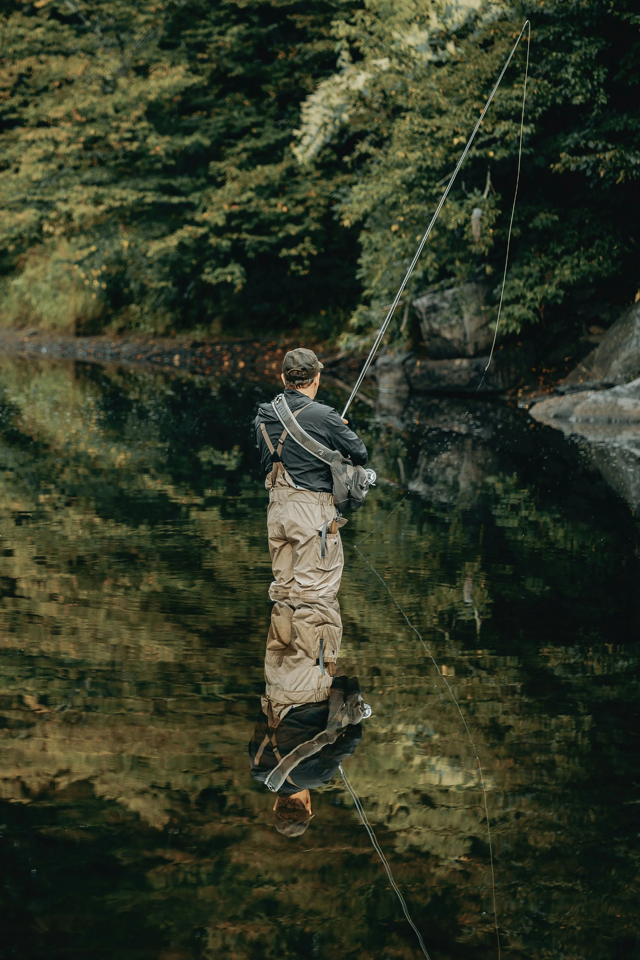 A person fishing in a river with a fishing rod, surrounded by dense green trees, and their reflection visible in the water.