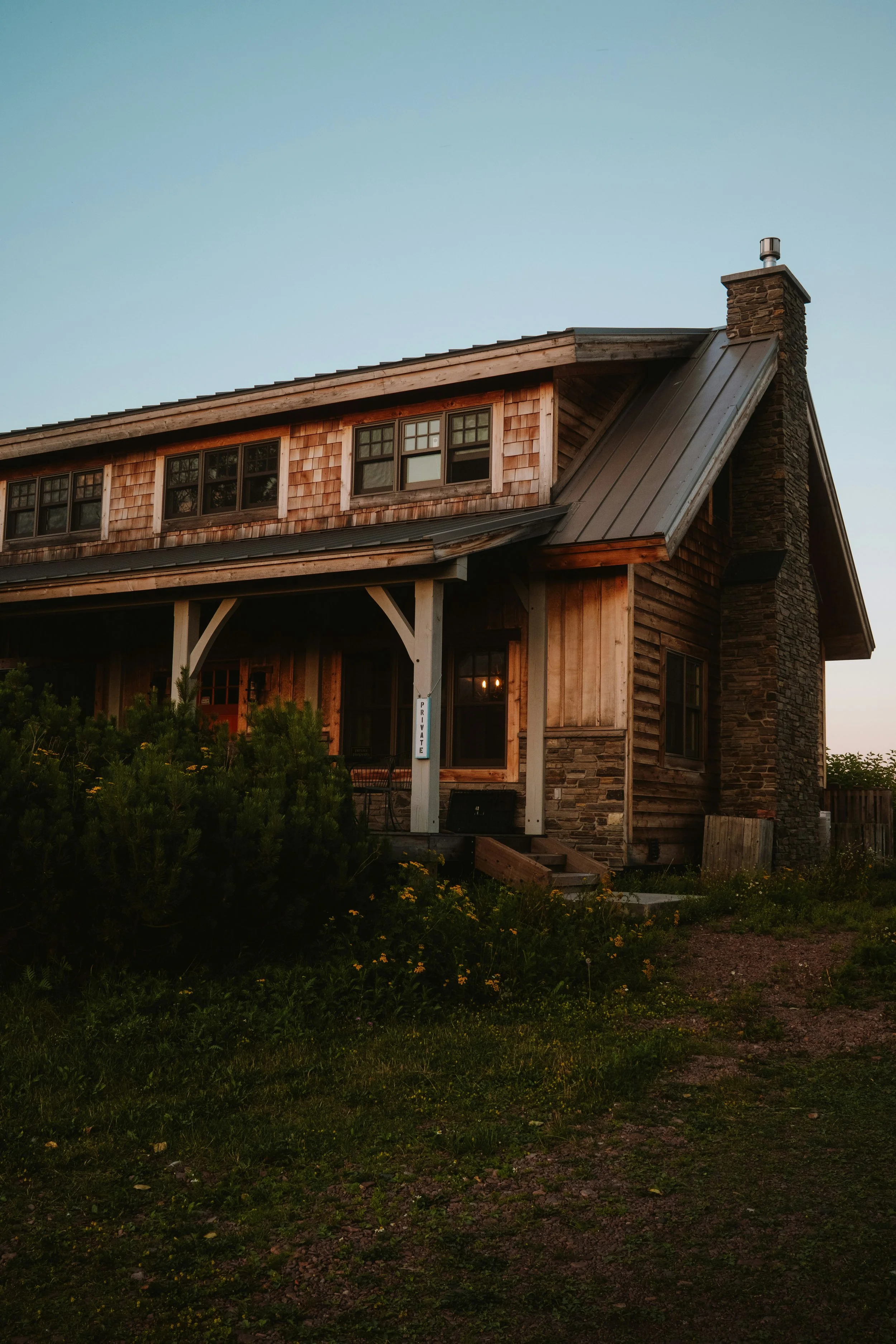 A two-story house with a wooden exterior, multiple windows, a steep metal roof, a stone chimney, and a front porch with seating, surrounded by greenery, at dusk.